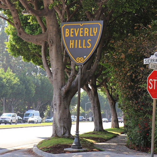 在一条绿树成荫的街角，矗立着一个“比佛利山庄”的标志，背景中有车辆，还有一个停车标志。