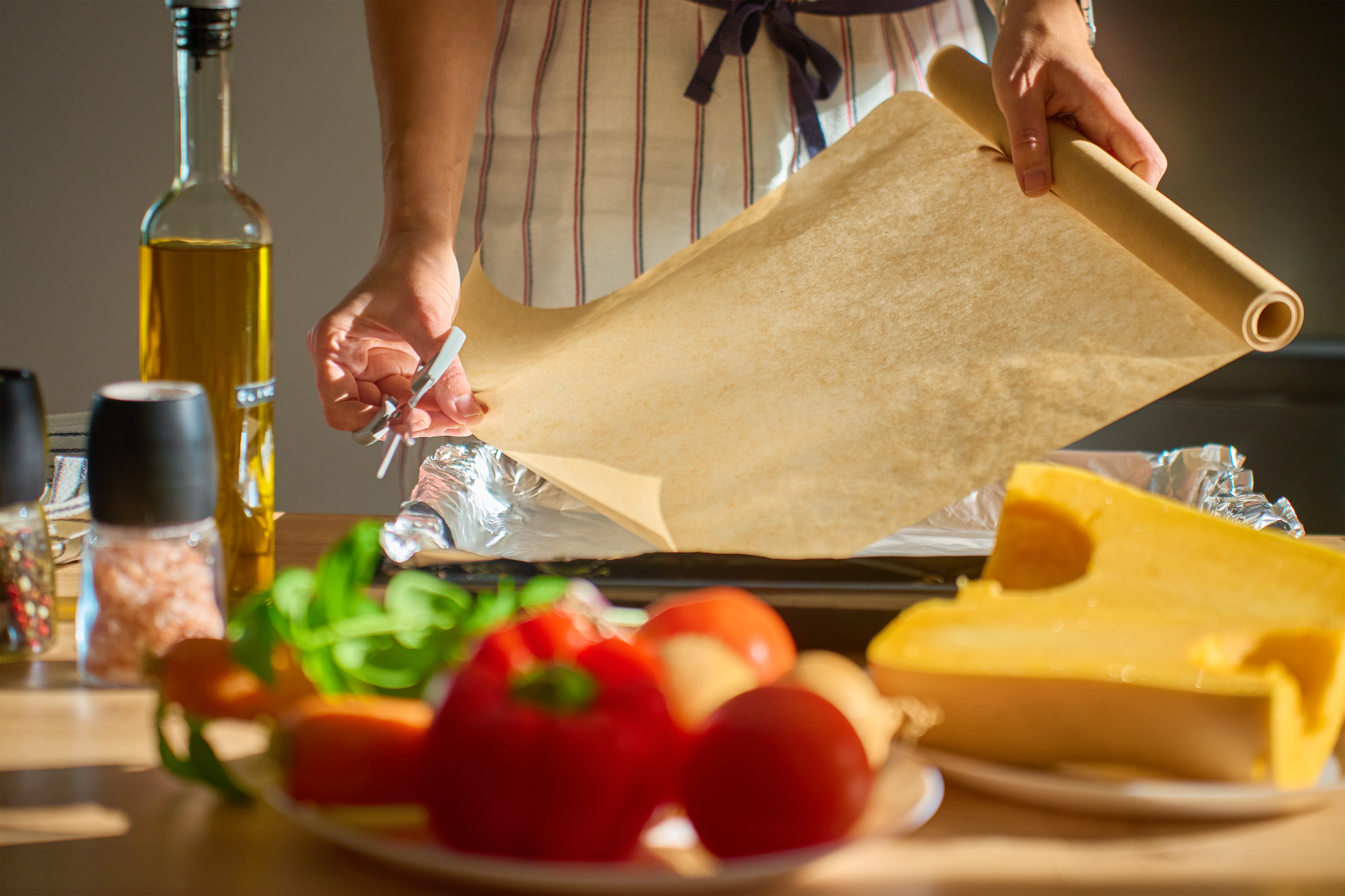 Person in apron placing baking paper on tray in sunlight kitchen with fresh vegetables, pumpkin, species and olive oil on table.
