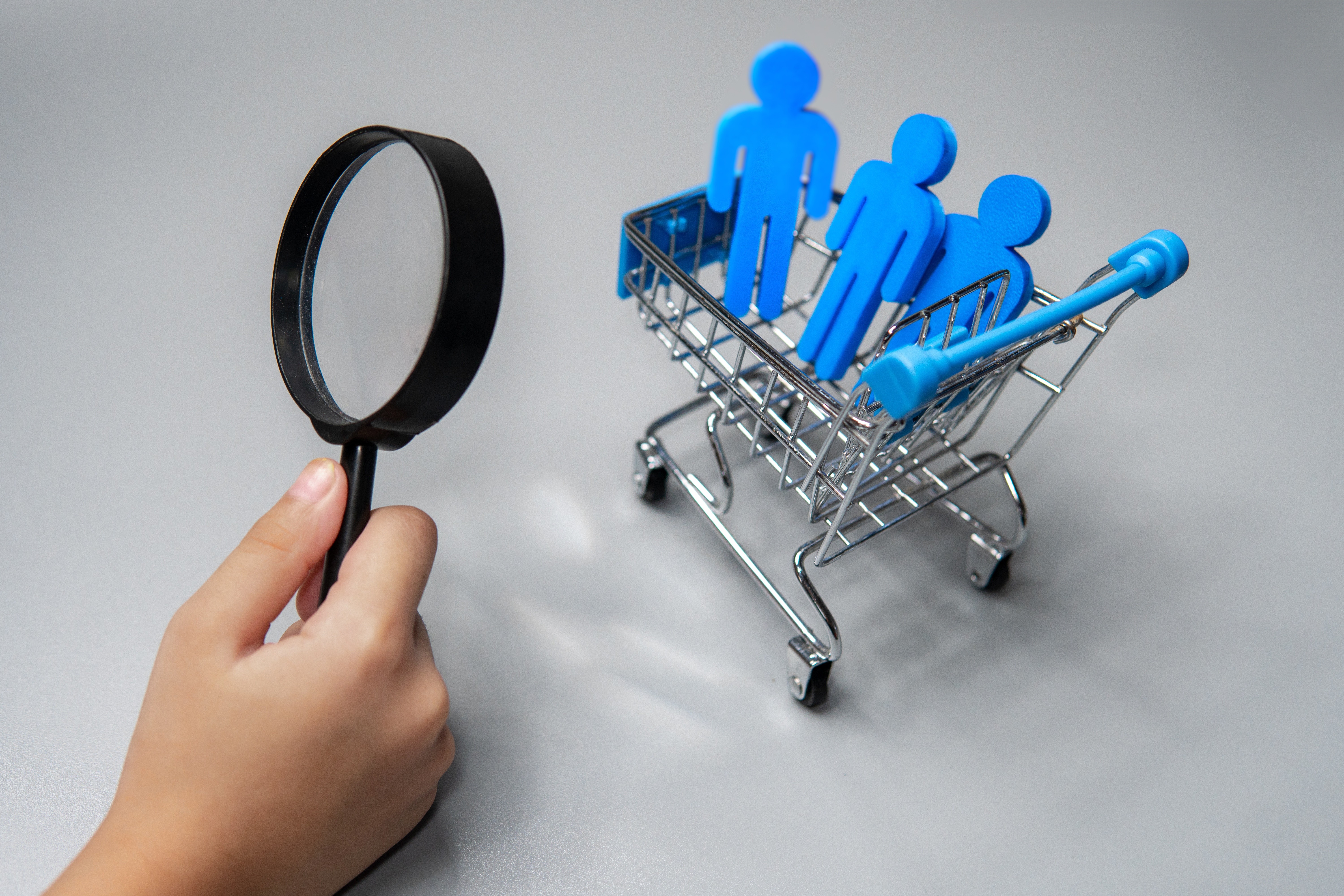 A hand holding a magnifying glass focusing on blue miniature human figures inside a miniature shopping cart. This conceptual image represents consumer market research, target audience analysis, and customer segmentation strategies. It effectively visualizes the process of identifying consumer behavior and buying patterns through detailed data analysis and retail market insights.