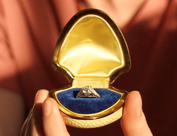 A diamond ring with a square-cut stone is nestled in a small, open gold ring box. The box has a rounded top and a textured base. The ring rests on a dark blue velvet cushion inside the box. The box is held between two fingers, partially visible in the frame. The background is softly blurred and appears to be a light peach or pinkish-orange color.