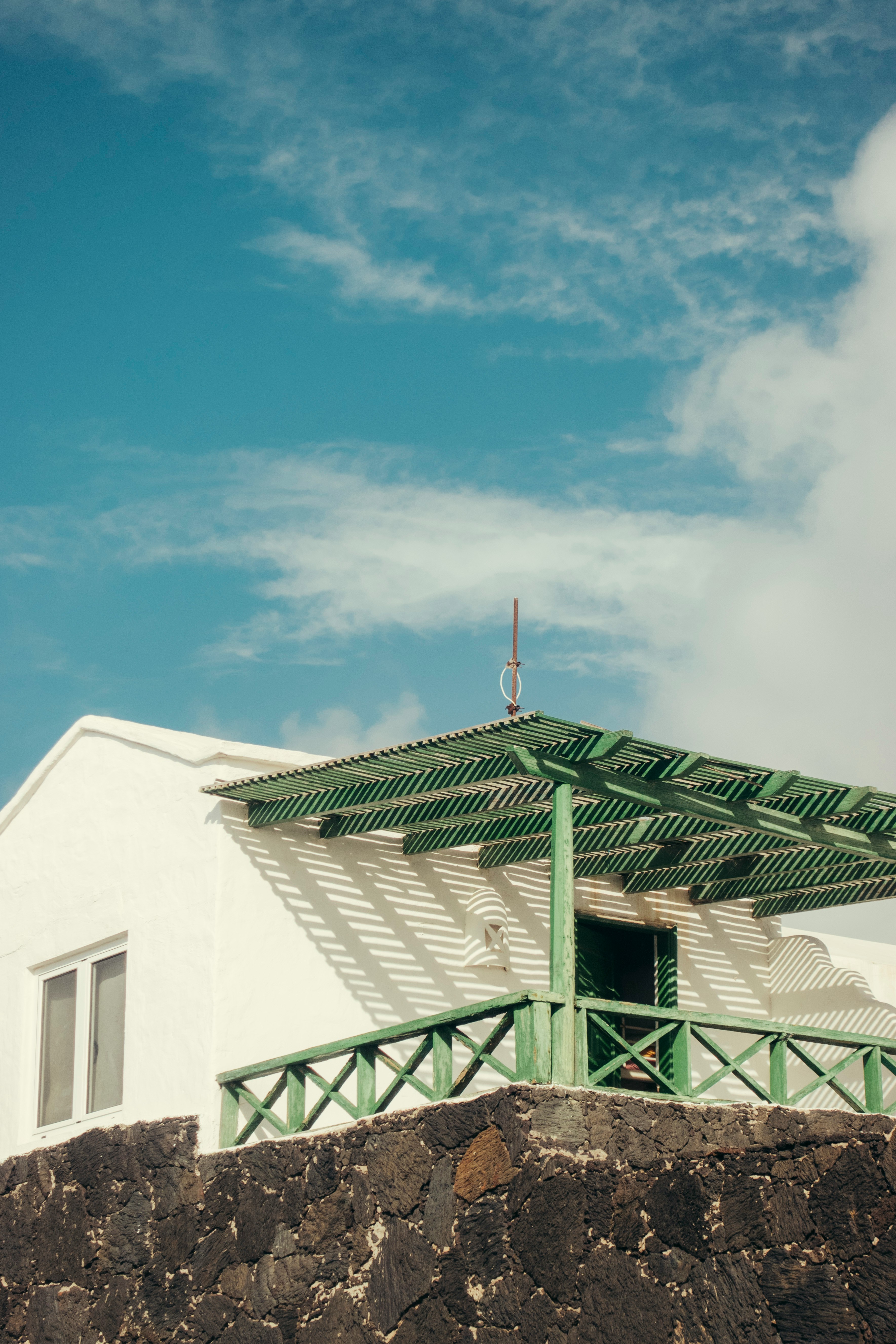 White house with green pergola on stone wall against blue sky.