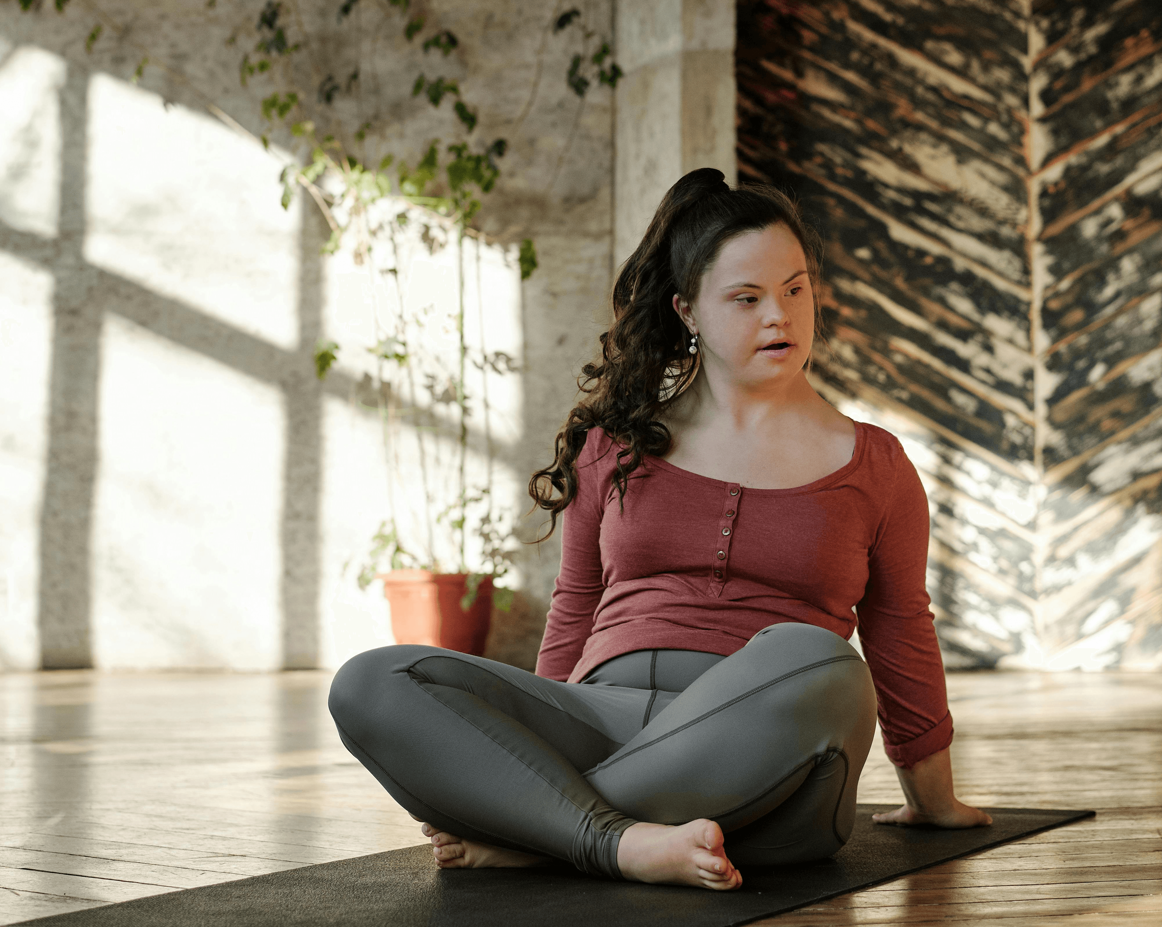 Woman with Down syndrome in a pink top and gray leggings sits cross-legged on a yoga mat in a sunlit room, exuding calm and focus. Indoor plants nearby.