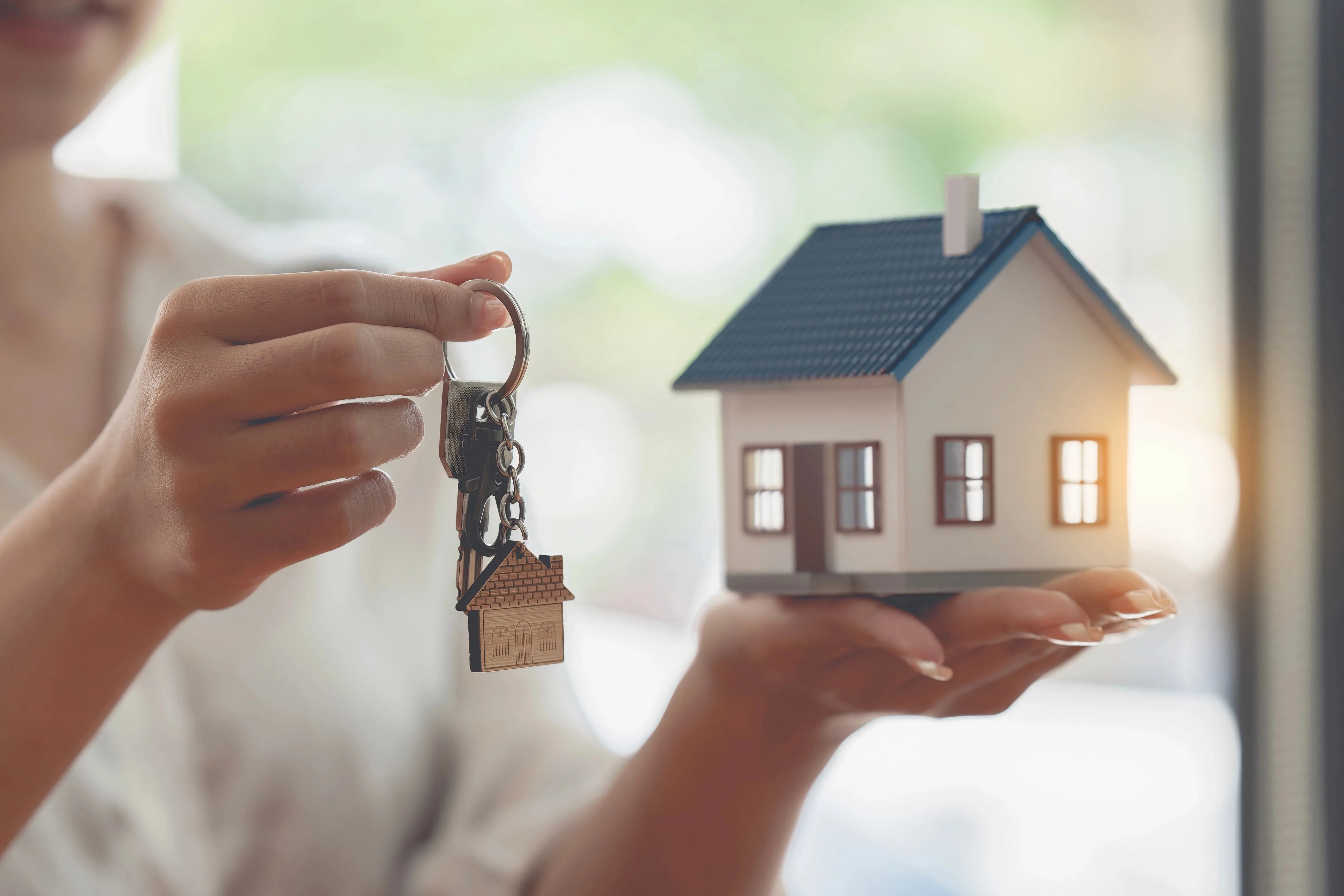 A person holding a house-shaped key charm in one hand while cradling a miniature model house in the other hand against a bright window background.