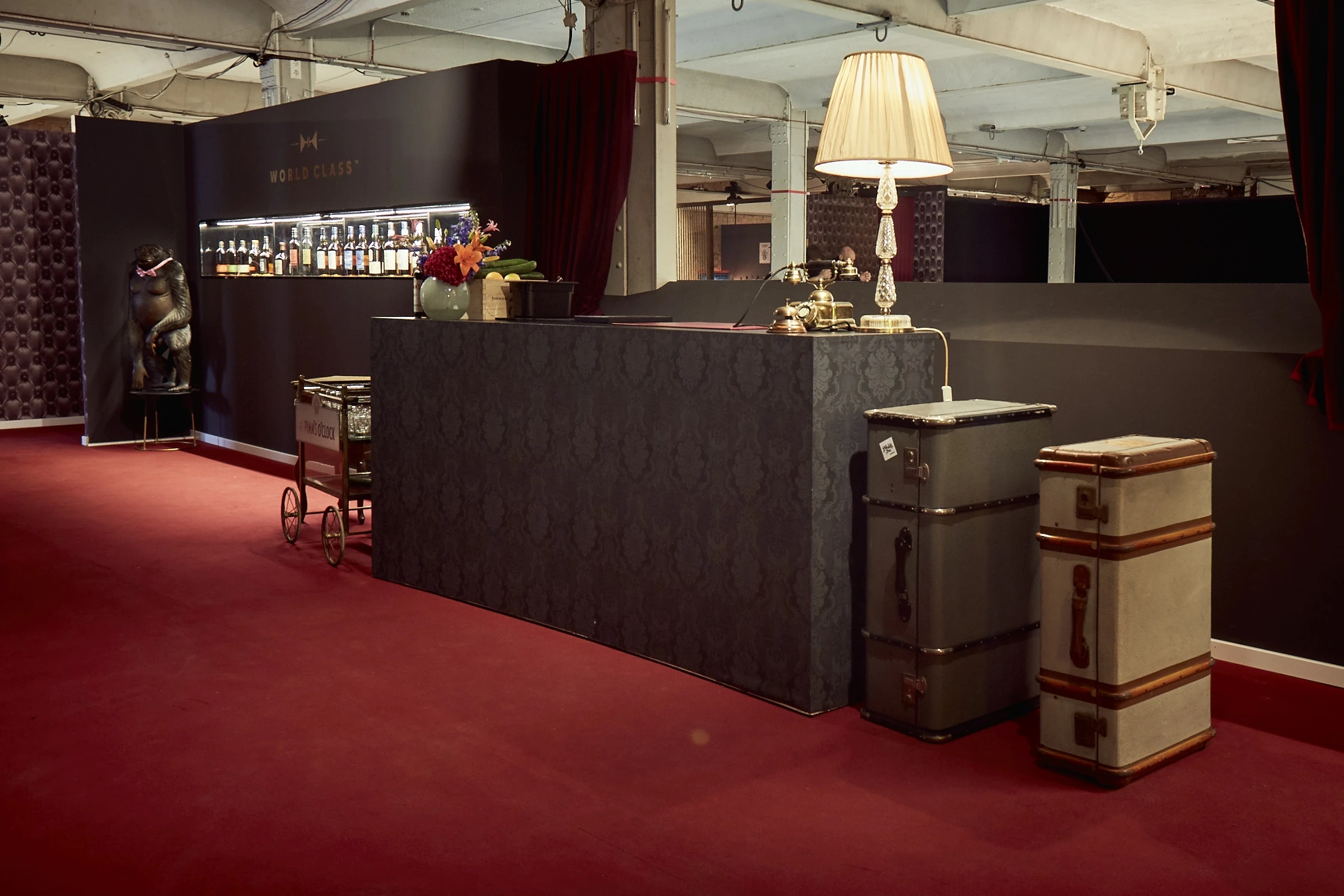 World Class bar at DIAGEO BCB17 with black damask-patterned counter, backlit bottle shelf, vintage trunks, a serving trolley and a fringed standing lamp on red carpet.