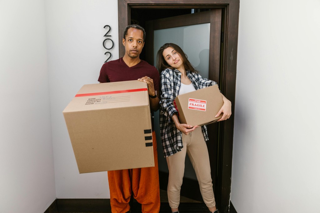 A man and woman holding large cardboard moving boxes (one marked "FRAGILE") in a doorway, representing inter-provincial relocation as newcomers move from expensive cities like Toronto or Vancouver to an affordable one like Calgary after successfully securing a job in the target province before moving.