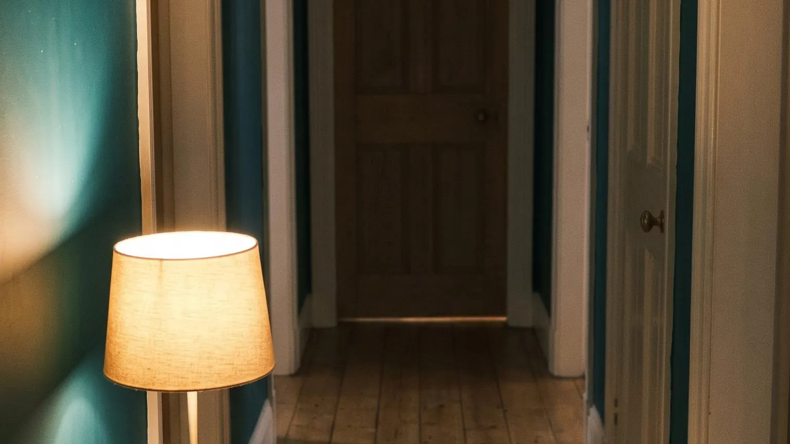 A hallway in the late evening lit by a table lamp, a deep teal wall and a closed door beyond