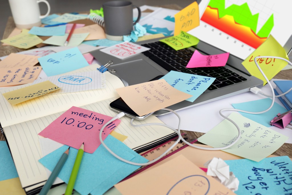 A cluttered desk covered in colorful sticky notes, loose papers, pens, and a laptop, with a coffee mug in the background, conveying overwhelm and disorganization.