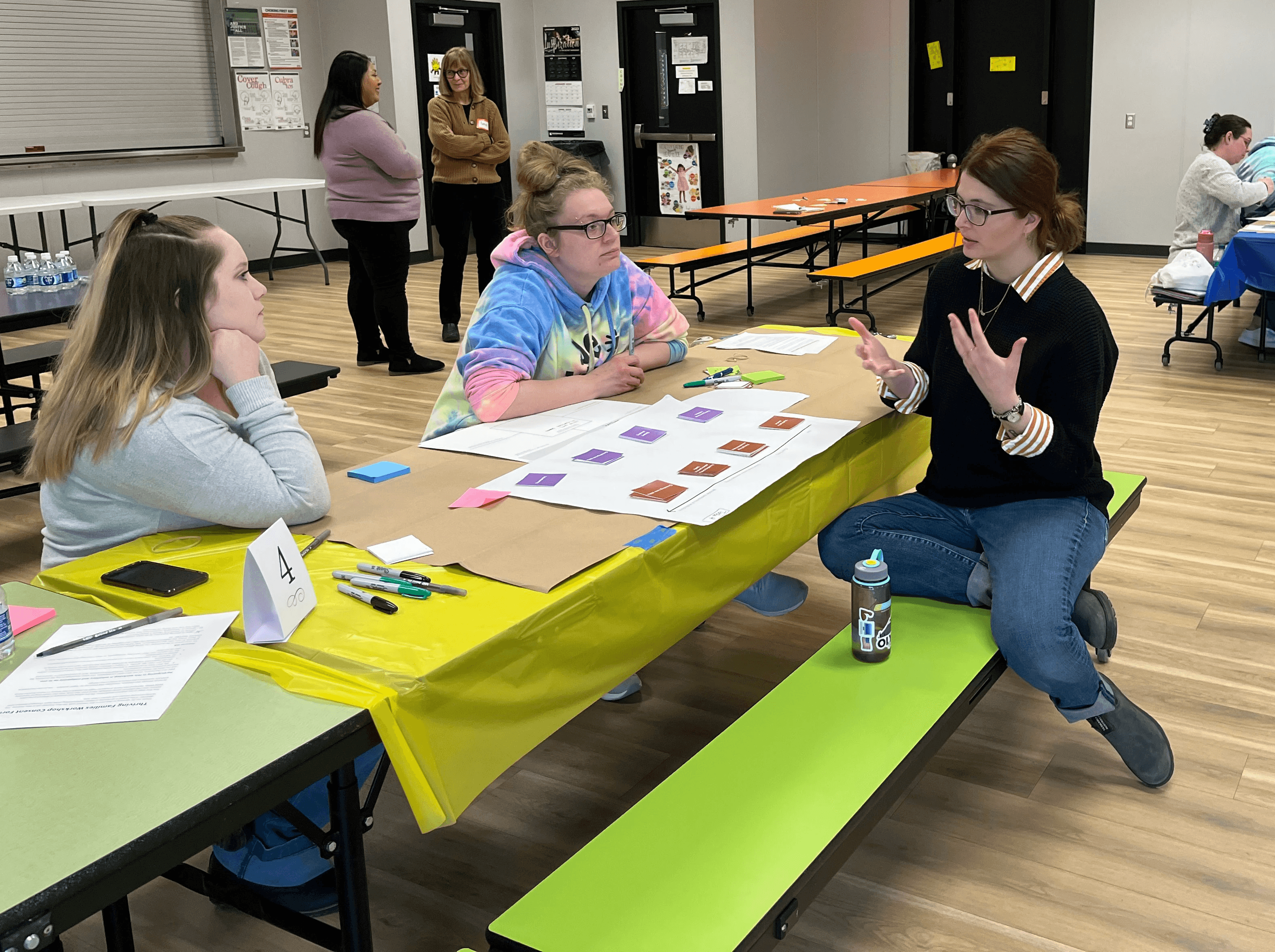 Three people in a workshop session, sitting around a table with post-it notes and papers around the table, with more people working in the background.