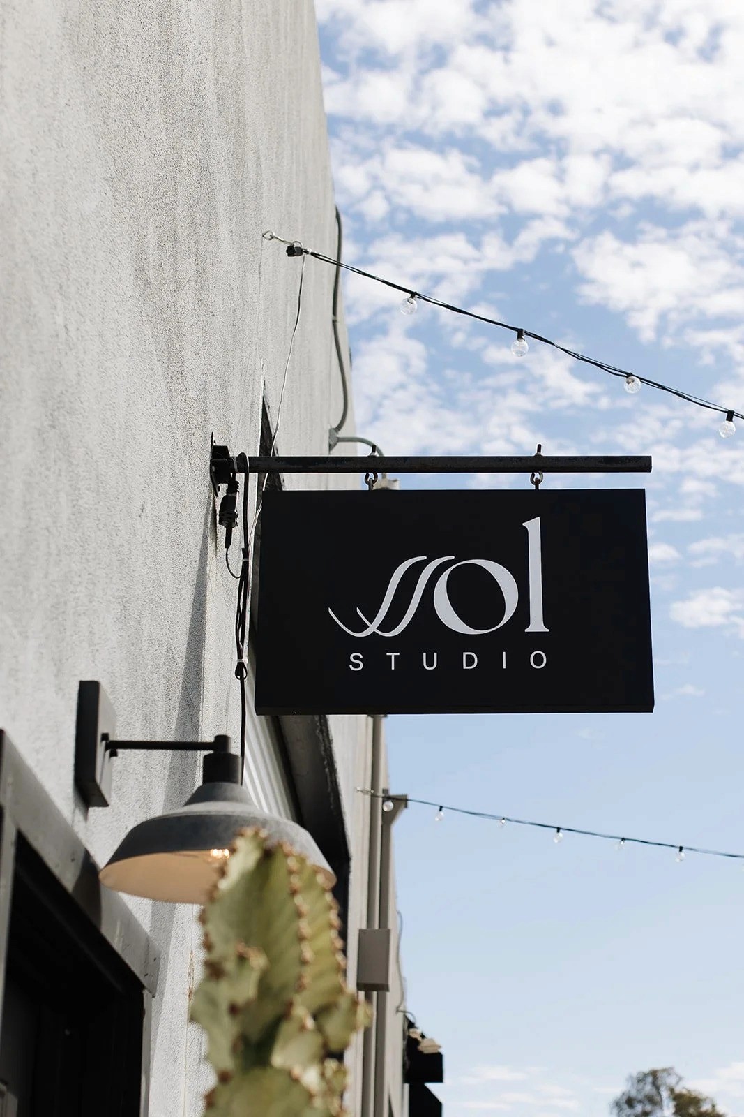 Black hanging storefront sign reading “sol studio” mounted on a metal bracket against a light stucco building, with string lights overhead and a blue sky with scattered clouds in the background.