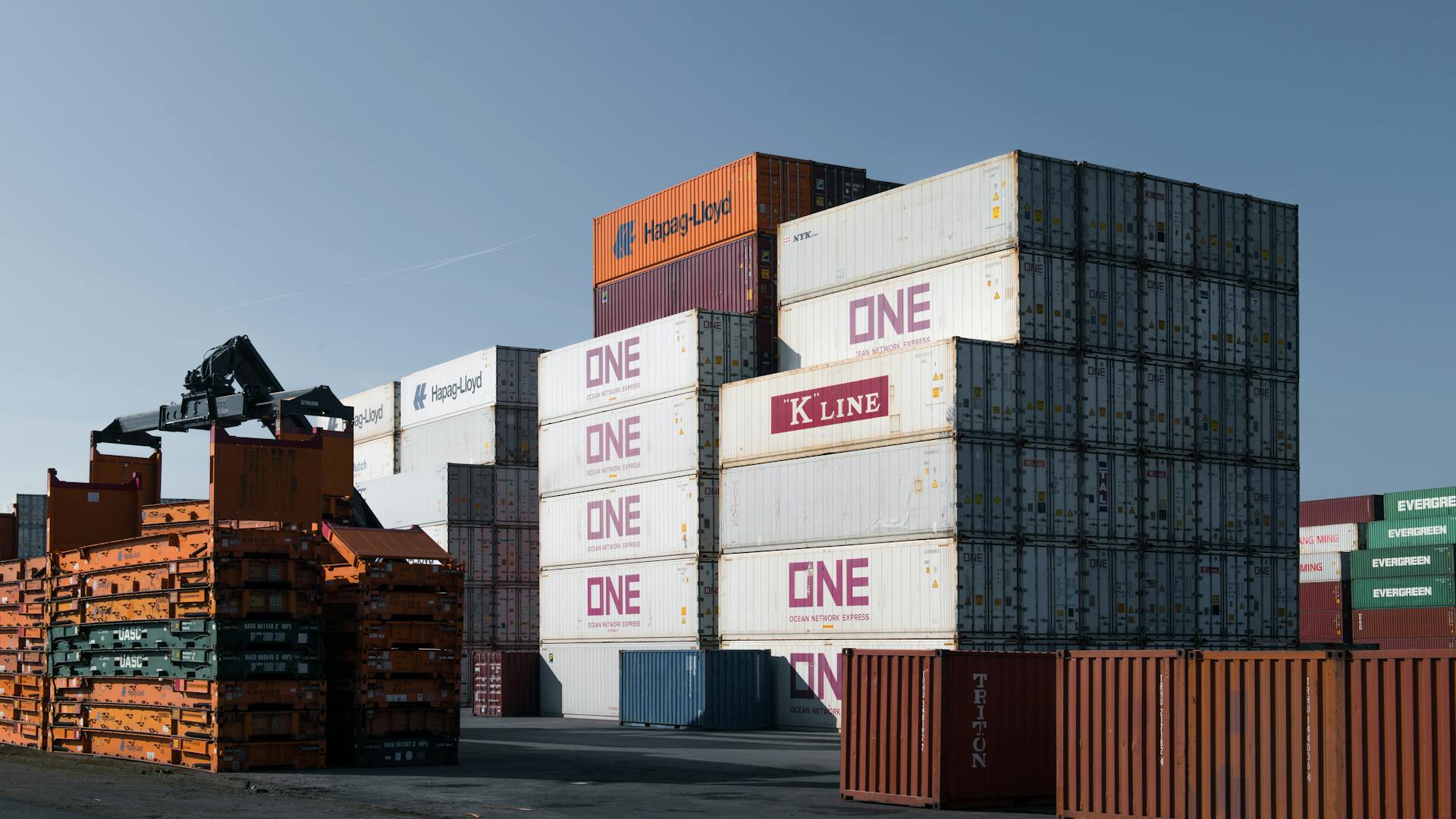 Stacked shipping containers at a port yard with a crane lifting cargo containers in the background.