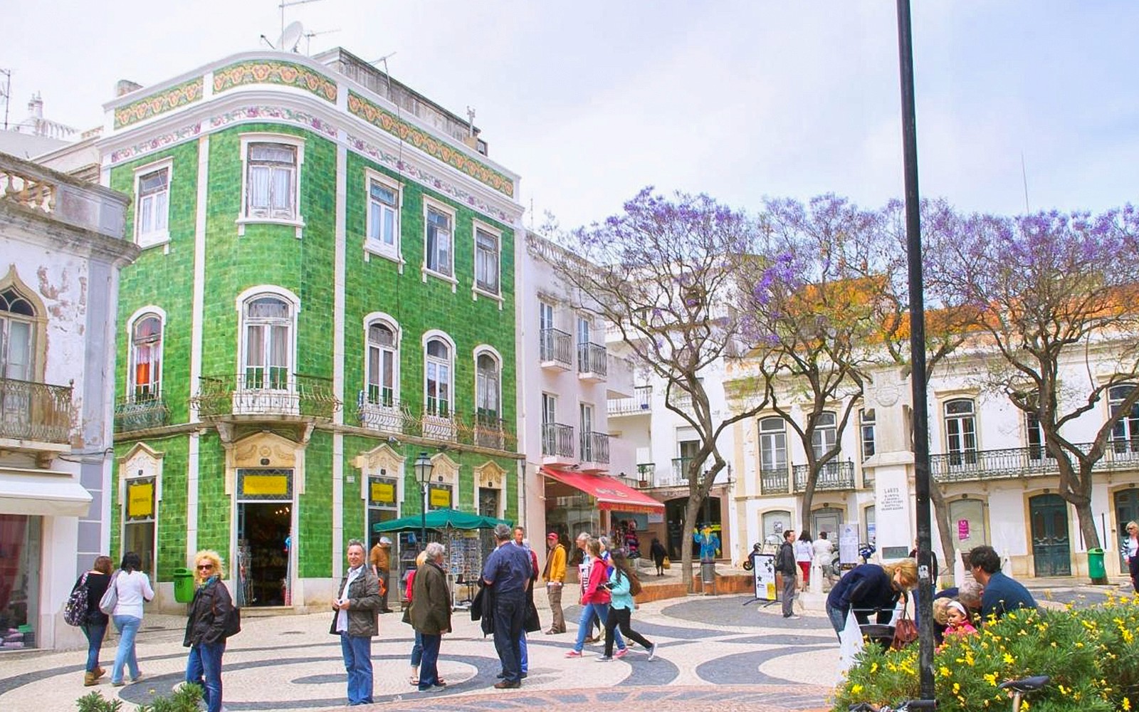 Pessoas caminhando em uma praça histórica com um edifício de azulejos verdes no Algarve Ocidental.