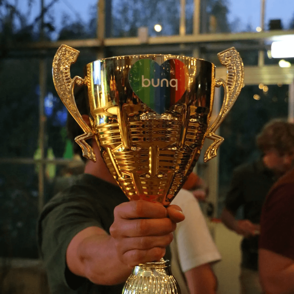 Een persoon die een gouden trofee vasthoudt met het bunq-logo hart in het midden, viert een overwinning in een helder verlichte binnenomgeving, omringd door mensen.