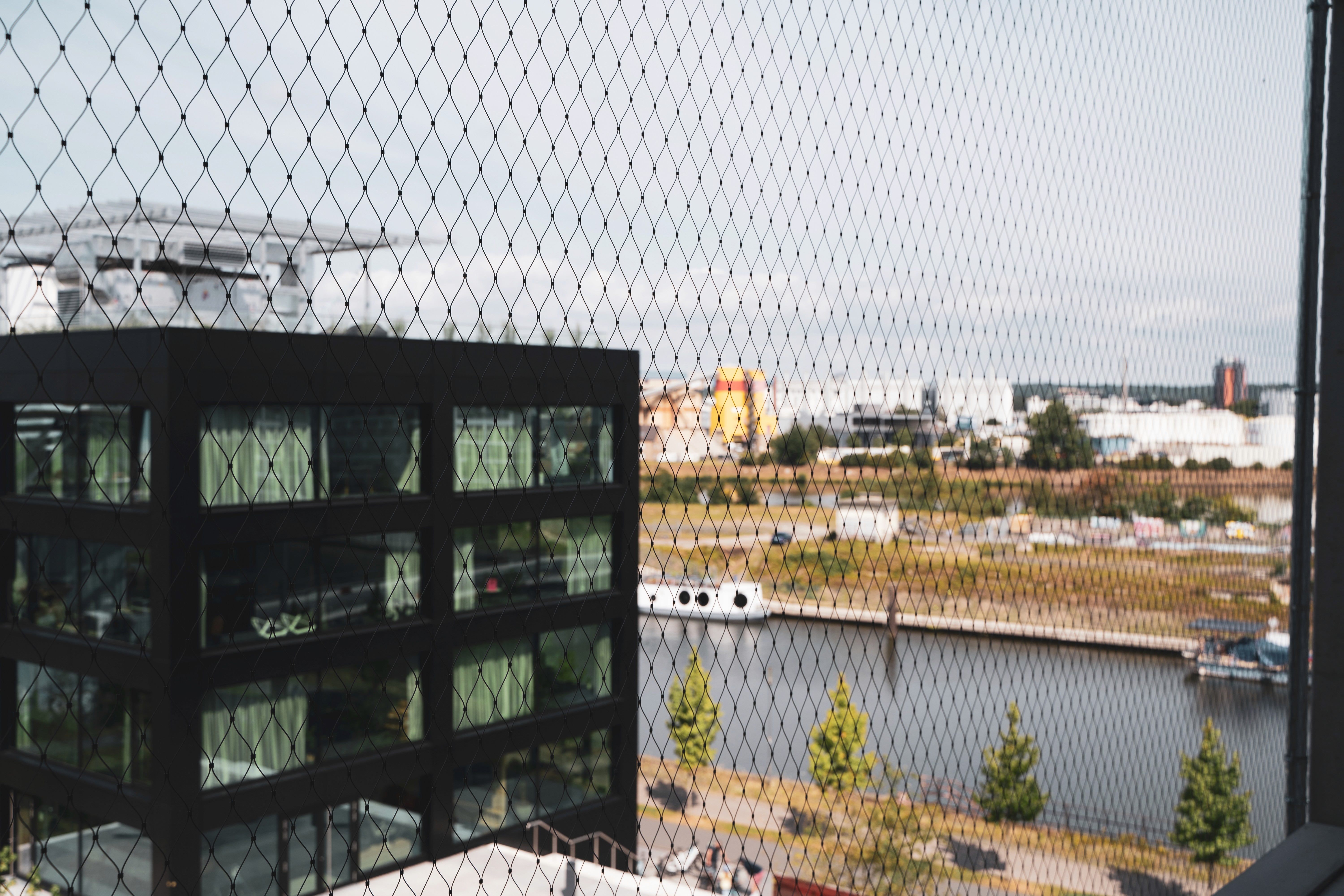 Blick durch einen Zaun auf den Hafen von Offenbach mit Wasser und modernen Gebäuden. Symbol für urbanes Arbeiten in inspirierender Umgebung.
