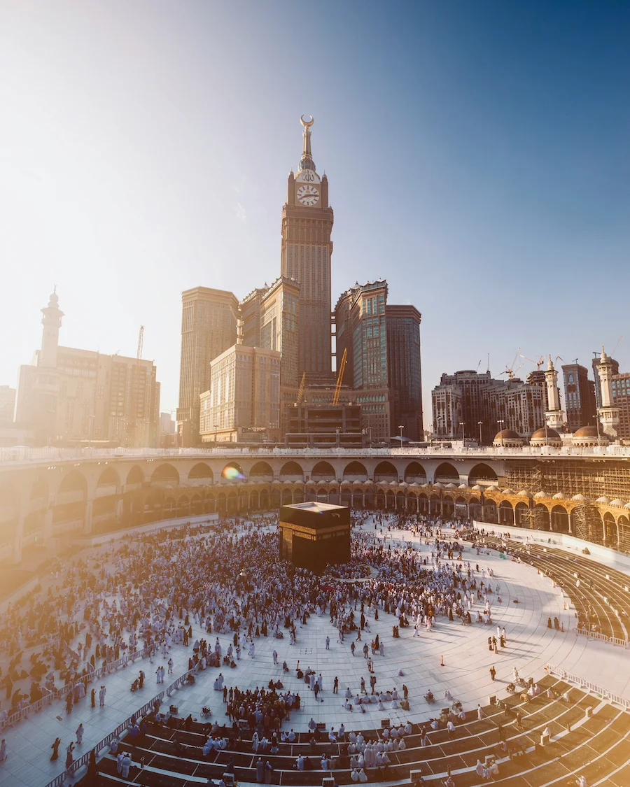 The Kaaba at Masjid al-Haram in Mecca with Abraj Al Bait Clock Tower behind and pilgrims gathered.
