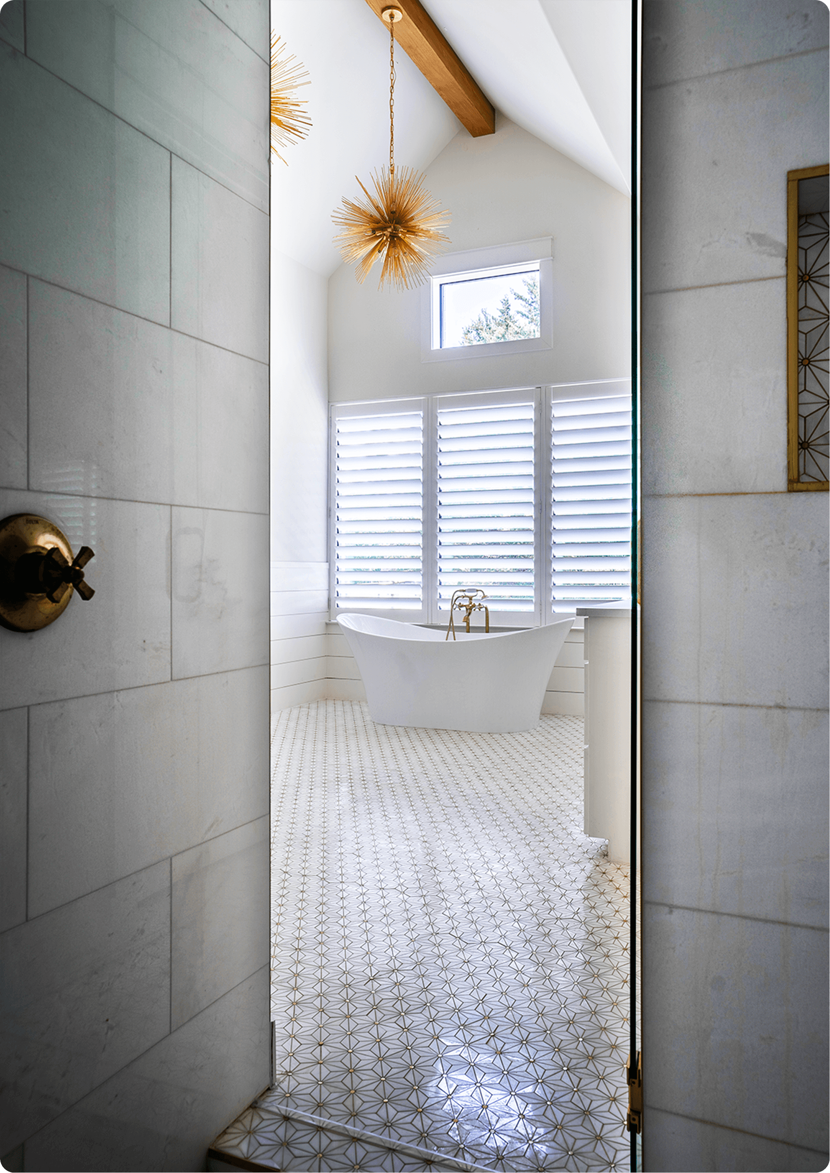 Modern bathroom with geometric patterned floor tile, white freestanding tub, brass fixtures, and white shutters