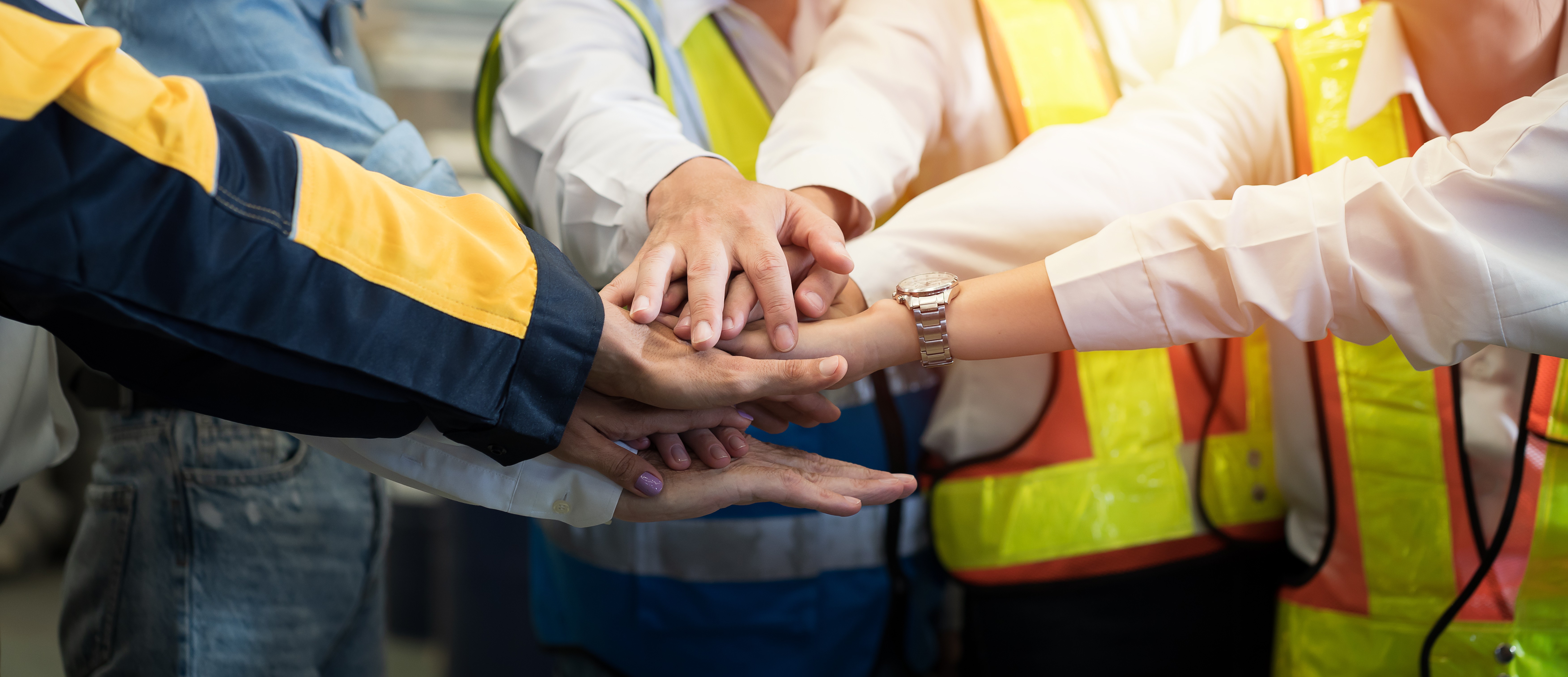 A group of professionals wearing safety vests and work uniforms stacking hands together in a teamwork gesture.