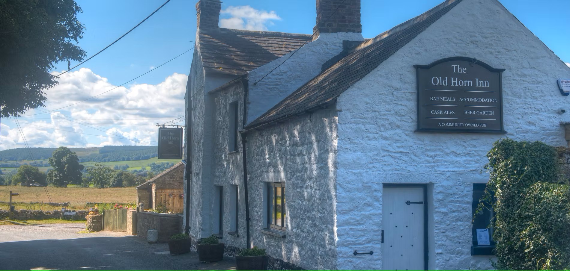 The Old Horn Inn, a whitewashed country pub with brick chimneys and a sign on its facade. The building features a white entrance door and is set in a rural location with open countryside visible in the background under blue skies.