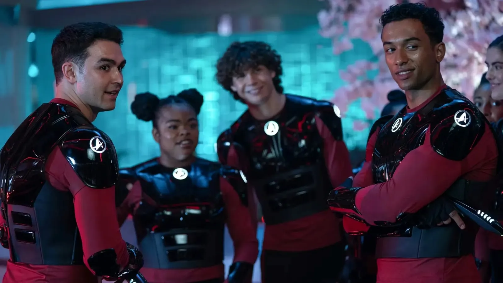 A group of people dressed in matching red and black futuristic suits gather together, smiling and engaging in conversation in a high-tech, illuminated environment.