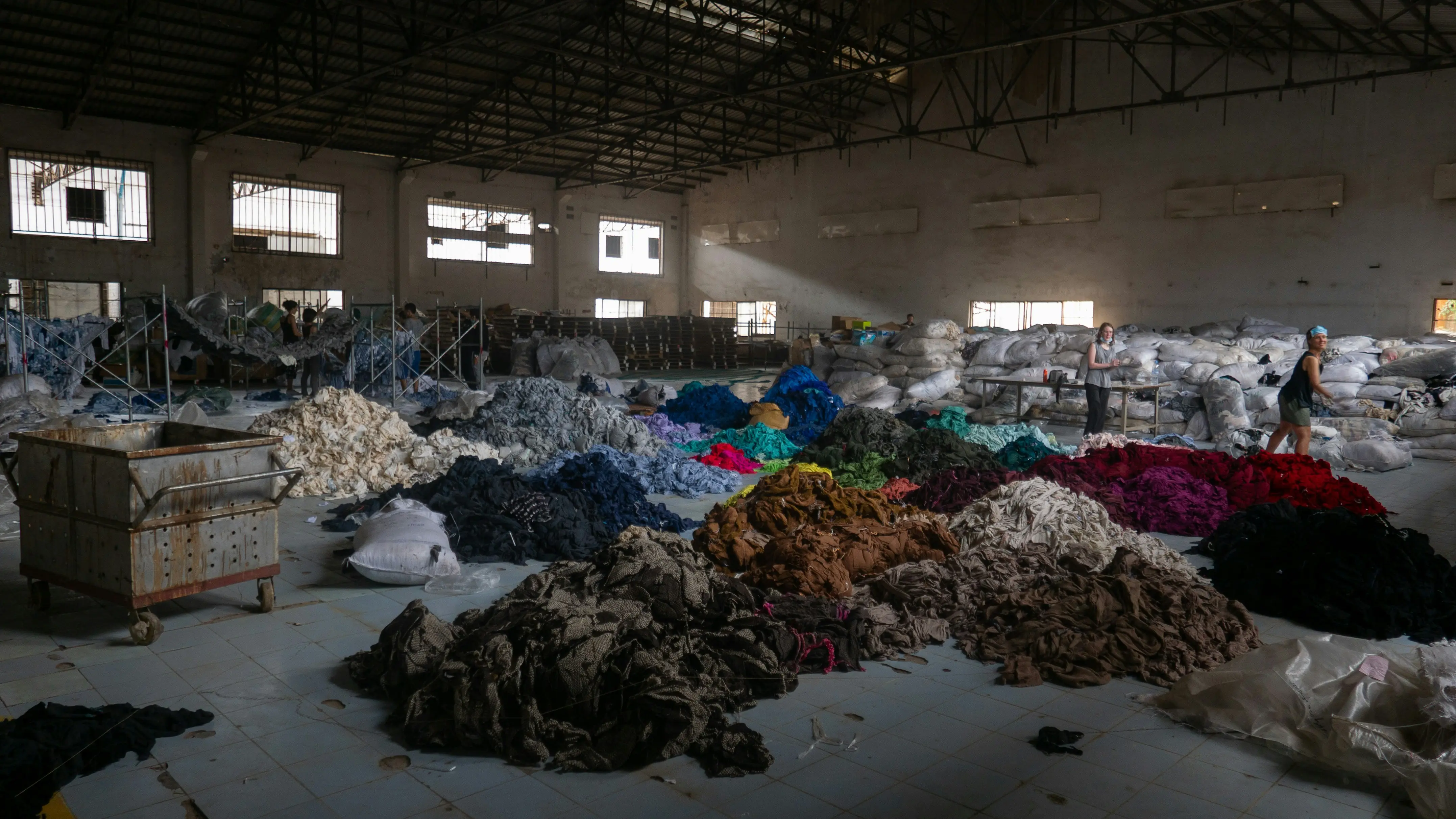 A large warehouse space filled with scattered piles of colorful fabric scraps and textile waste, with workers organizing materials in the background.