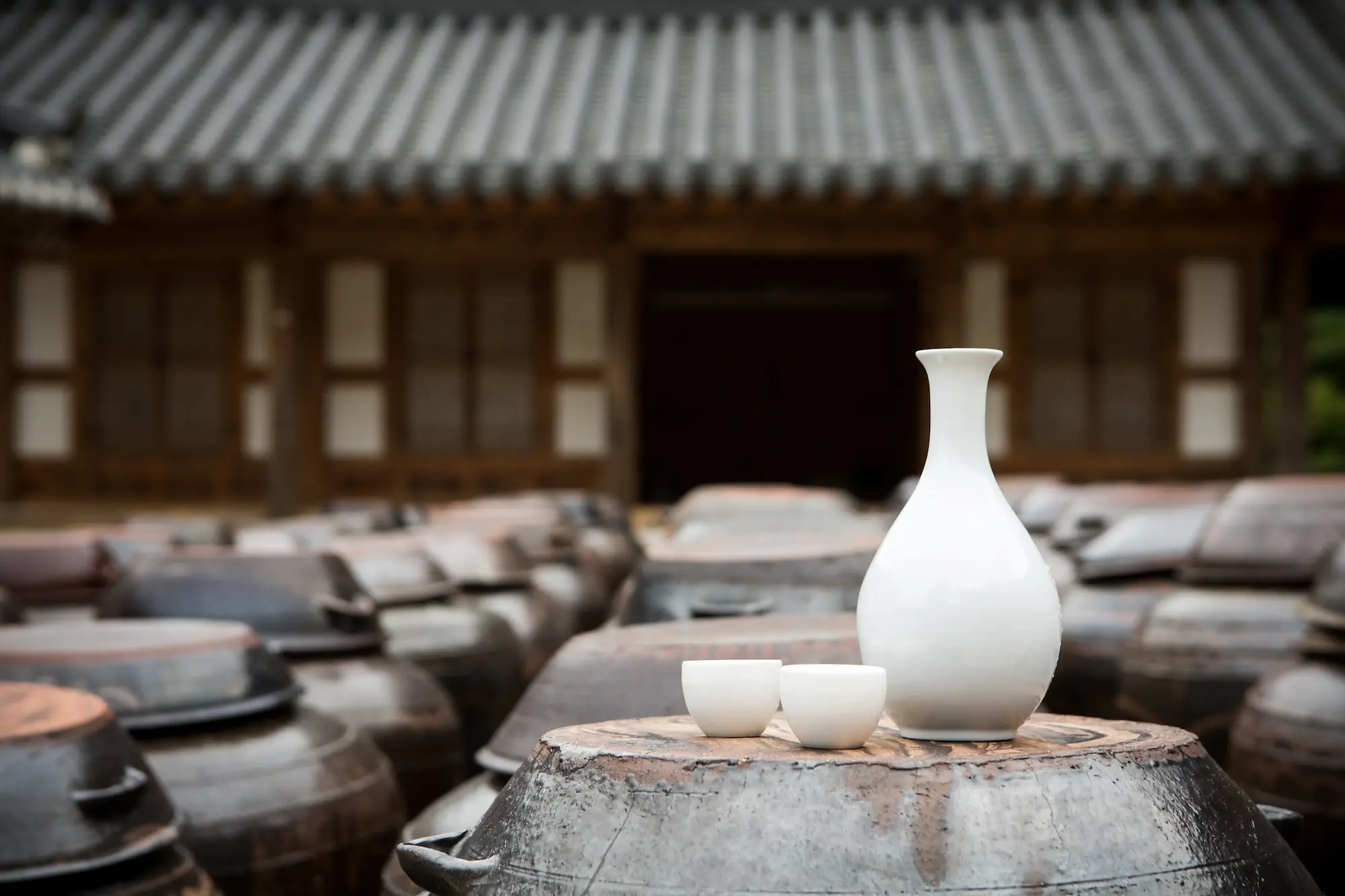 A white porcelain bottle set, consisting of a bottle and two cups, is elegantly displayed on a traditional wooden barrel in front of a classic Korean hanok building.