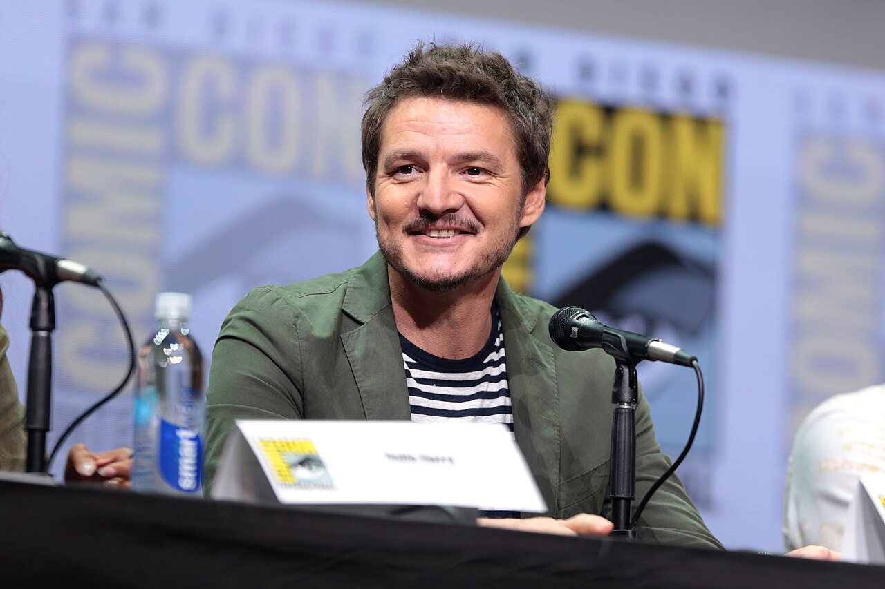 Pedro Pascal in a green jacket and striped shirt sits at a panel discussion, speaking into a microphone. A name card is partially visible in front of him. A bottle of water and blurred Comic-Con signage are in the background.