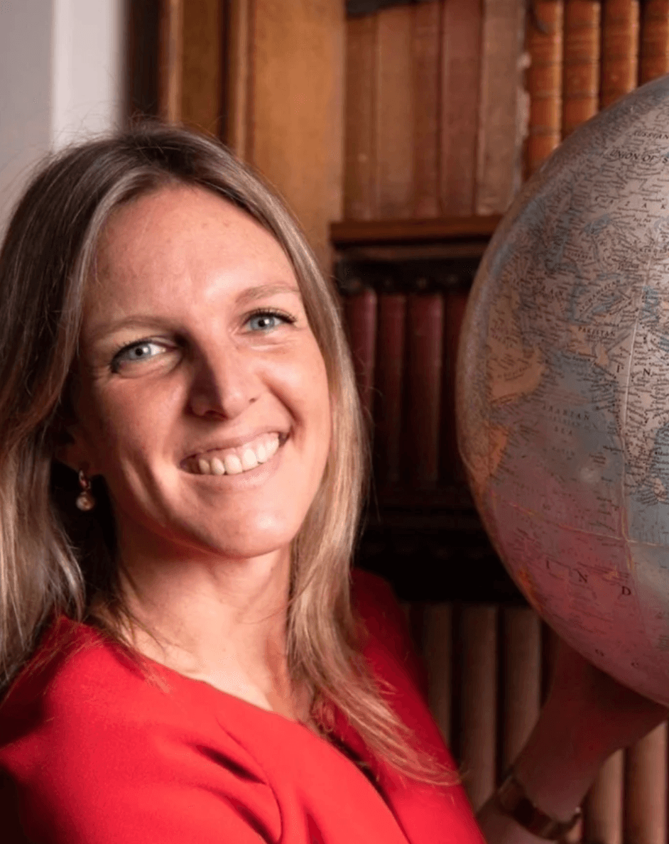 Person wearing a blue puffer jacket and red backpack standing in front of a light blue building on a city street.