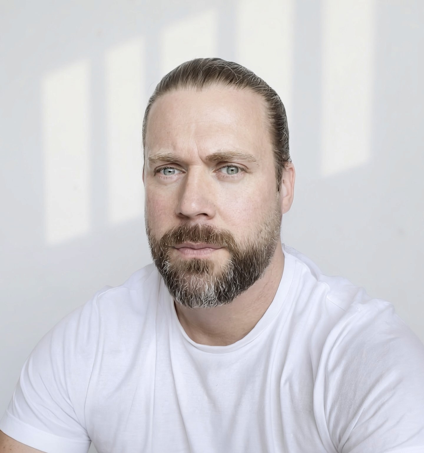 Casual studio portrait of a bearded man with curly hair in a white shirt, arms crossed, on a light gray background.