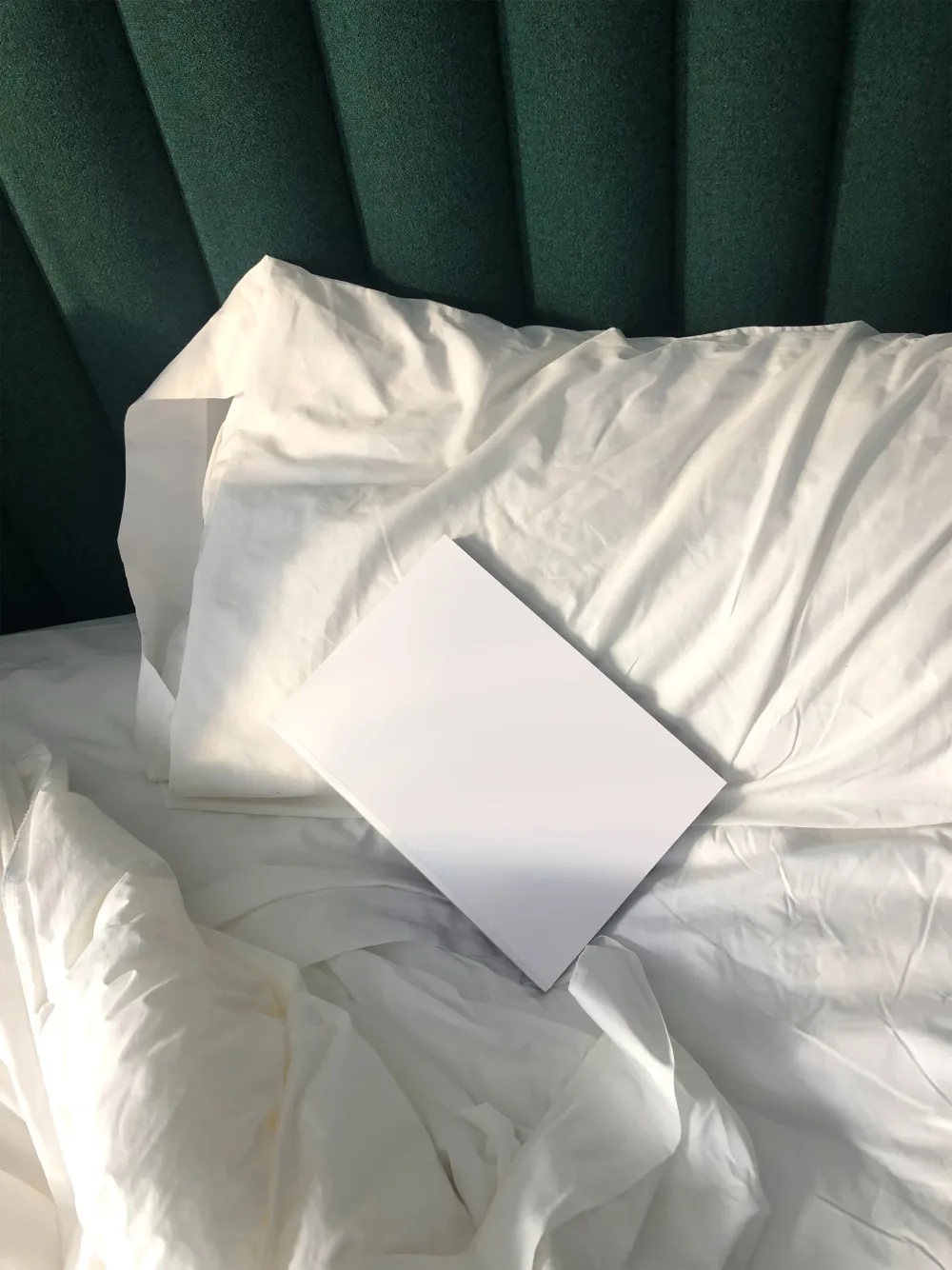A minimalistic hardcover book mockup placed on a crumpled white bedsheet, set against a green upholstered headboard. The book features bold black geometric patterns on a white cover. Soft natural light enhances the cozy ambiance.