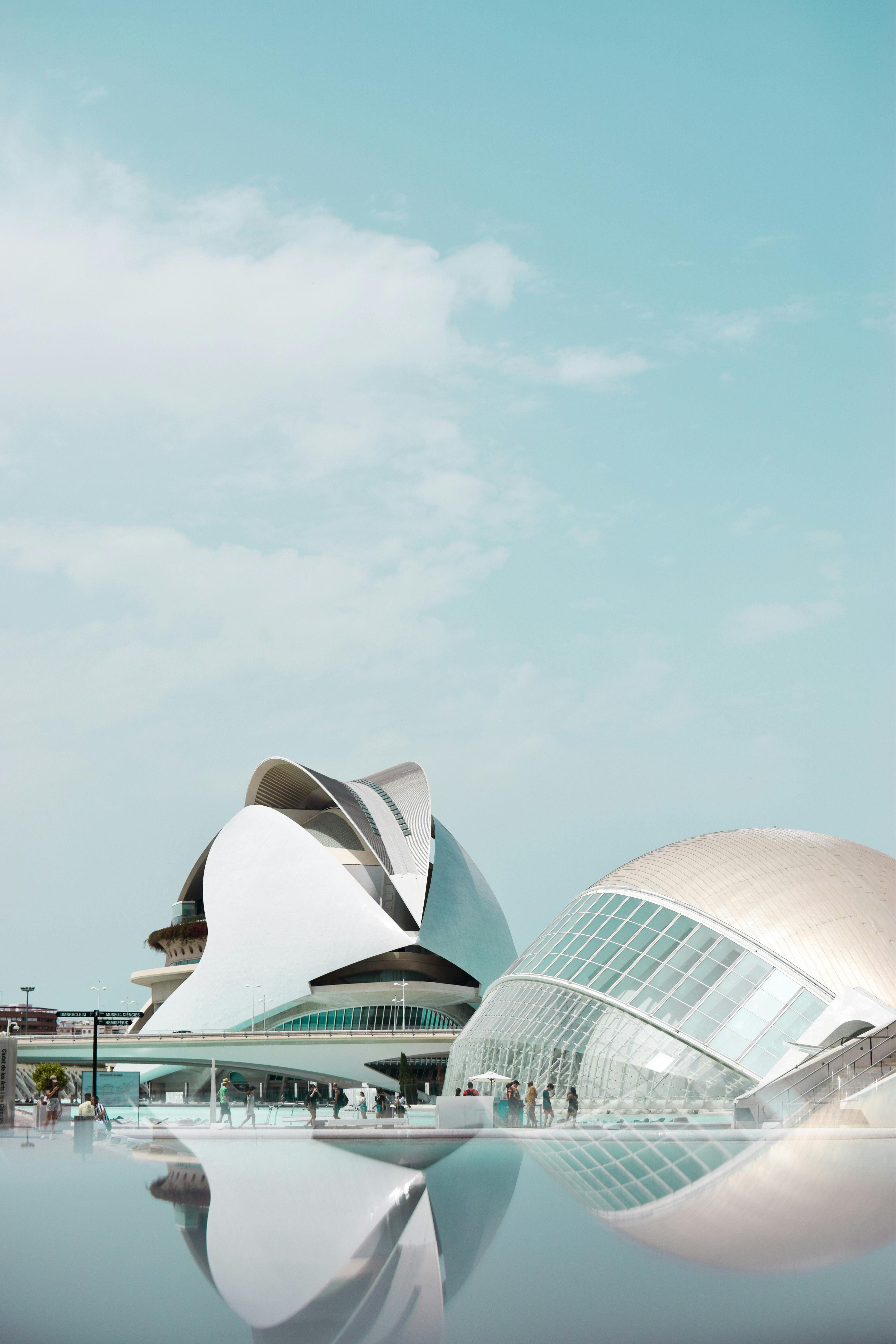 A stylish, modern lounge area featuring pastel green sofas, tan chairs, round wooden tables, and ceiling plants, with large windows and sheer curtains.
