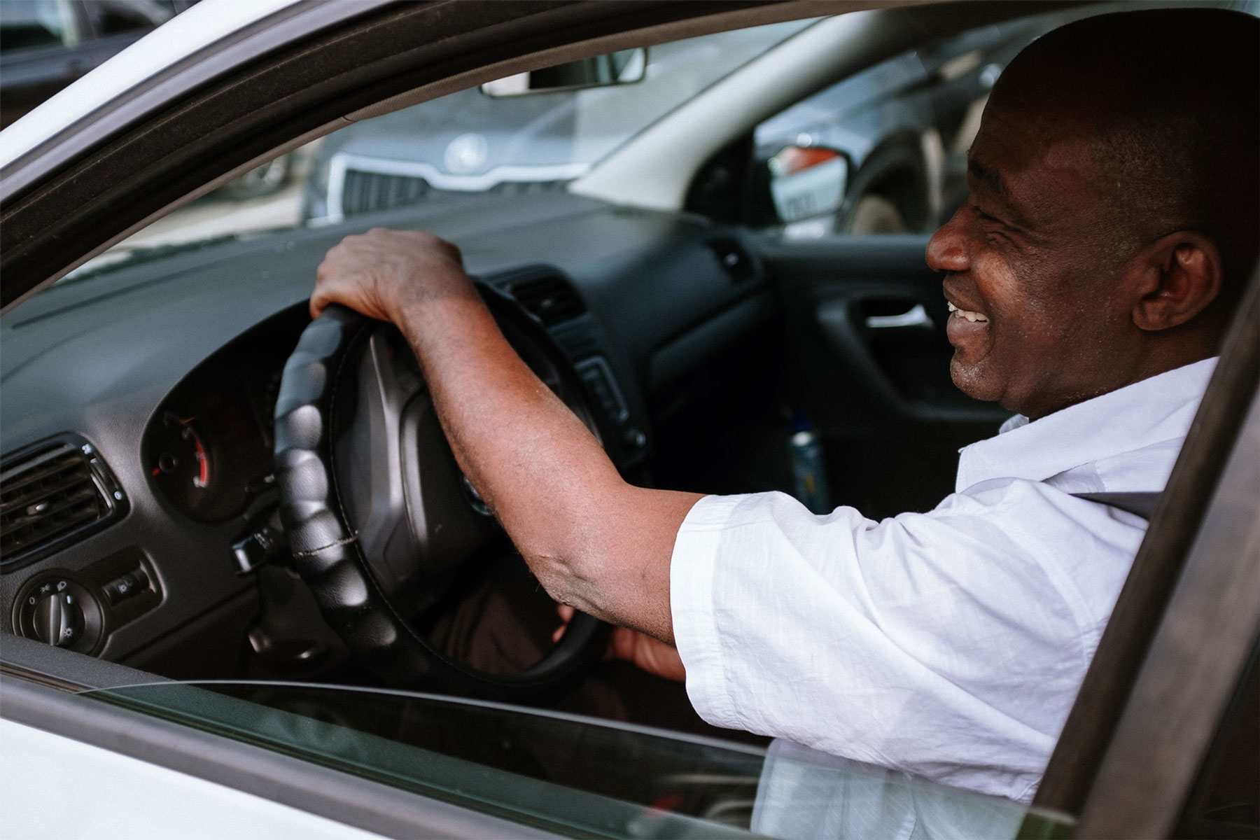 A smiling man in a driver's seat, gripping the steering wheel with a relaxed posture.