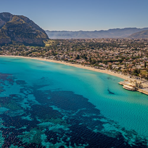 Aerial view of a coastal city with turquoise water, sandy beach, numerous buildings, and mountains in the background.