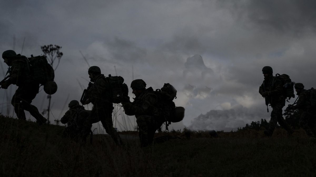 Black and white photo of 4 military personal tactically moving on flat terrain with clouded skies in background