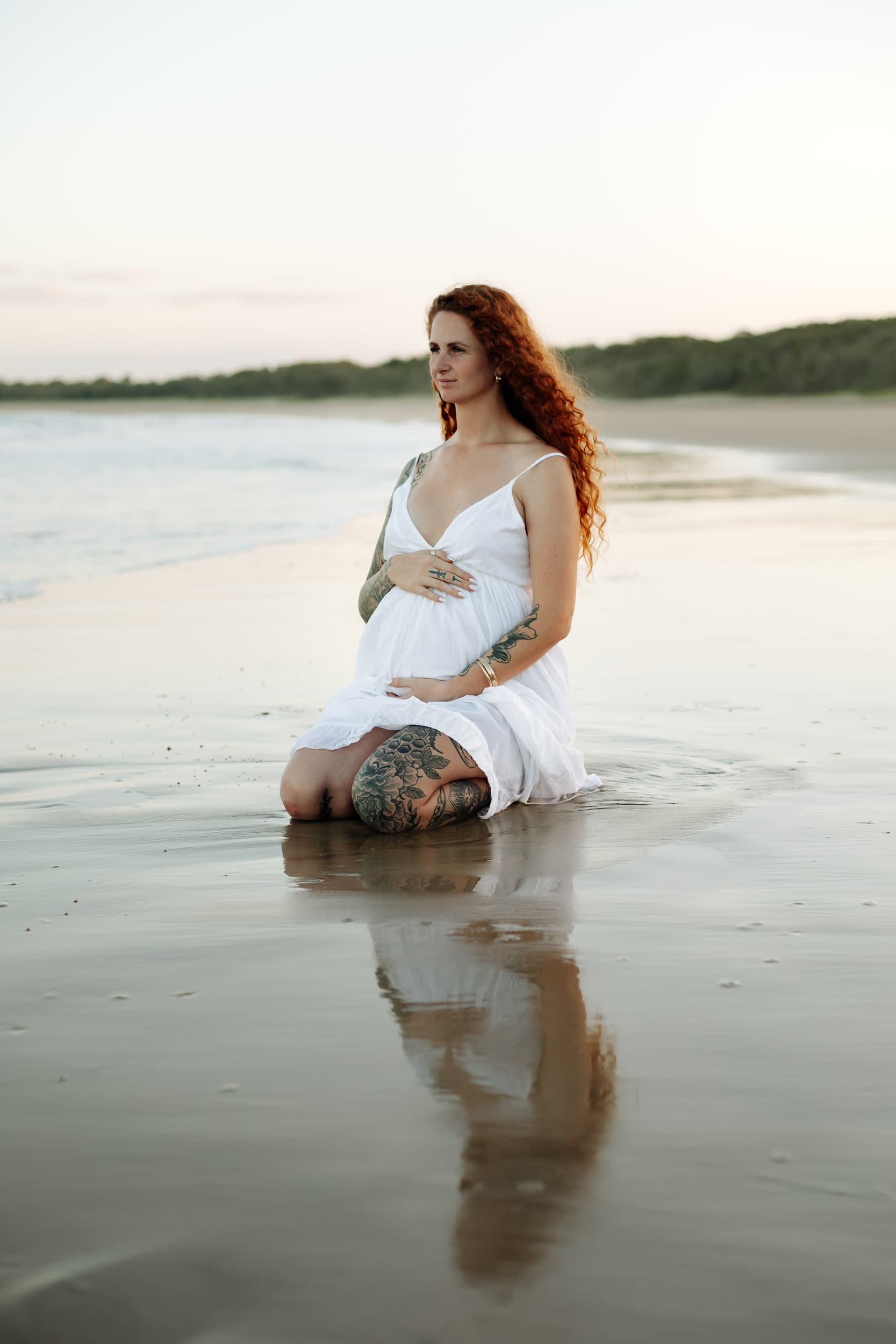 Solo maternity portrait of mother-to-be sitting in wet sand as the sun sets over the Mackay coastline.