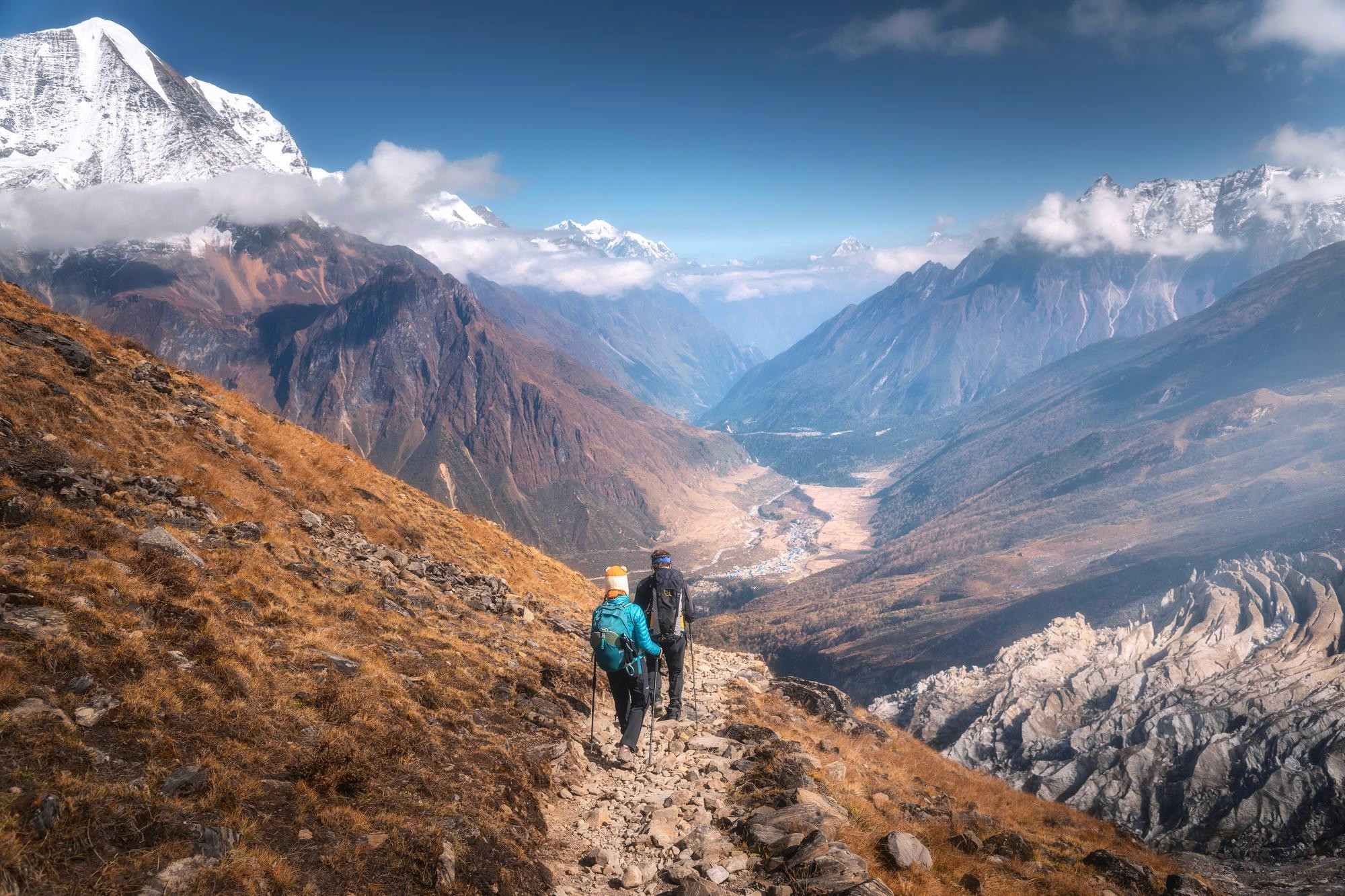 A man in a bright orange raincoat helps a woman in a yellow jacket step across large rocks on the shore of a calm mountain lake.