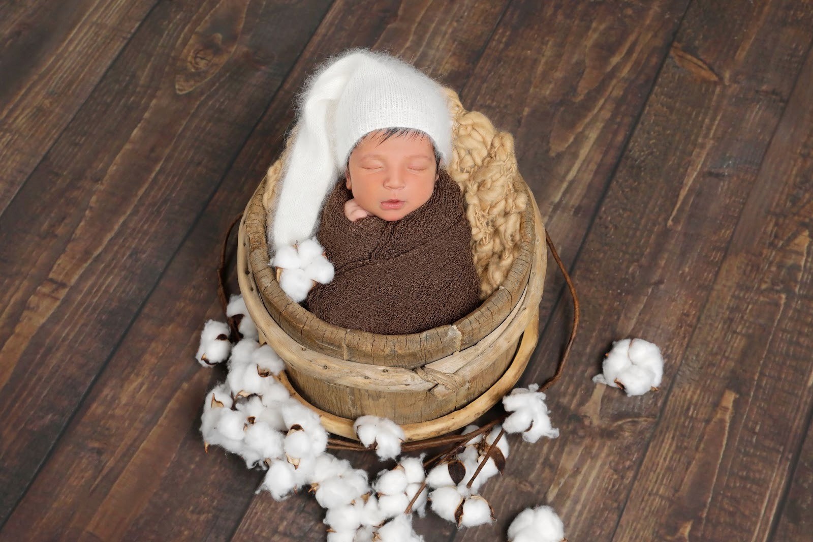 Swaddled baby wearing a white knit long-tail hat resting in a rustic wooden bucket.