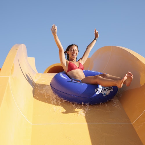 A woman in a red bikini enjoys a ride on a blue inner tube down a bright yellow water slide under a clear blue sky.