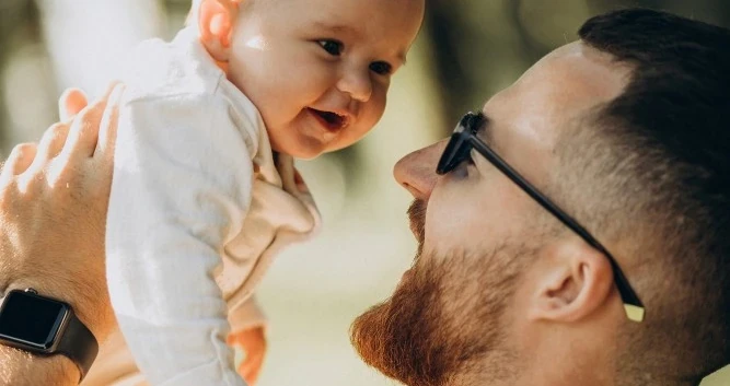 Homem de óculos erguendo um bebê sorridente ao ar livre, os dois se olhando