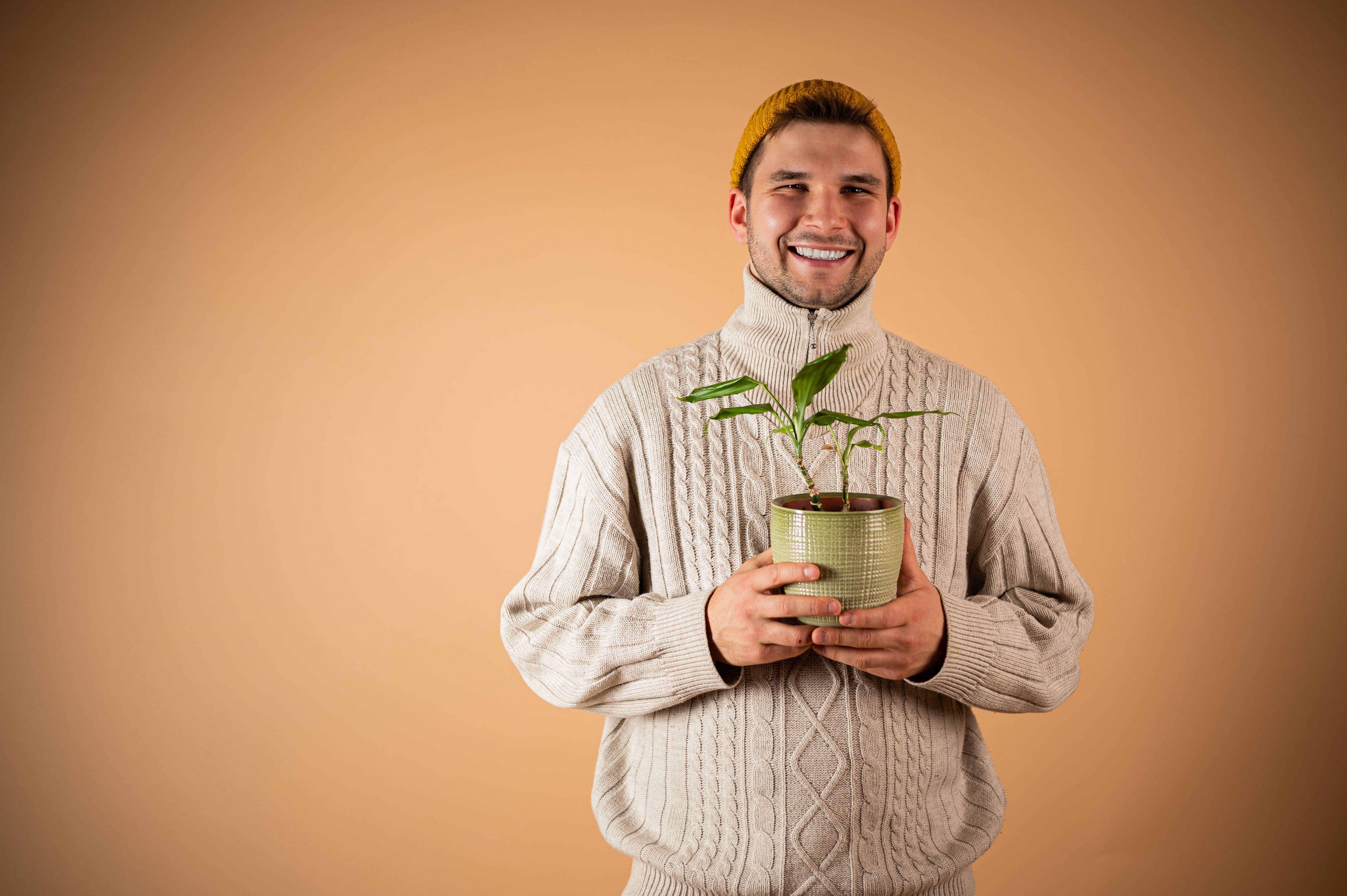 Ein lächelnder Mann in einem beigen Zopfmuster-Pullover hält behutsam eine kleine Topfpflanze in den Händen; der Hintergrund ist einfarbig orange-braun.