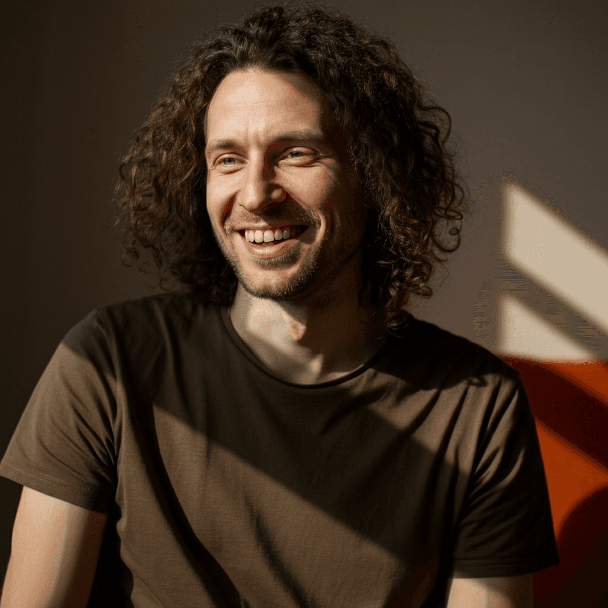 A smiling man with curly hair sits in soft sunlight, wearing a brown T-shirt. Shadows and warm tones create a relaxed, joyful atmosphere.