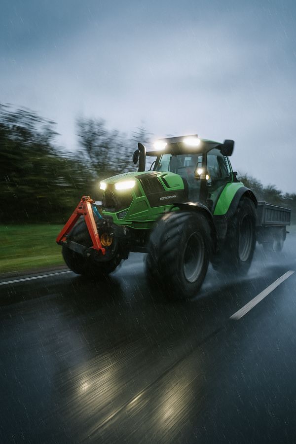 Ein moderner, grüner Deutz-Fahr Traktor mit eingeschalteten Scheinwerfern und Anhänger während der Fahrt im Einsatz für Erdbau-Projekte, Gartengestaltung Klingenstein.