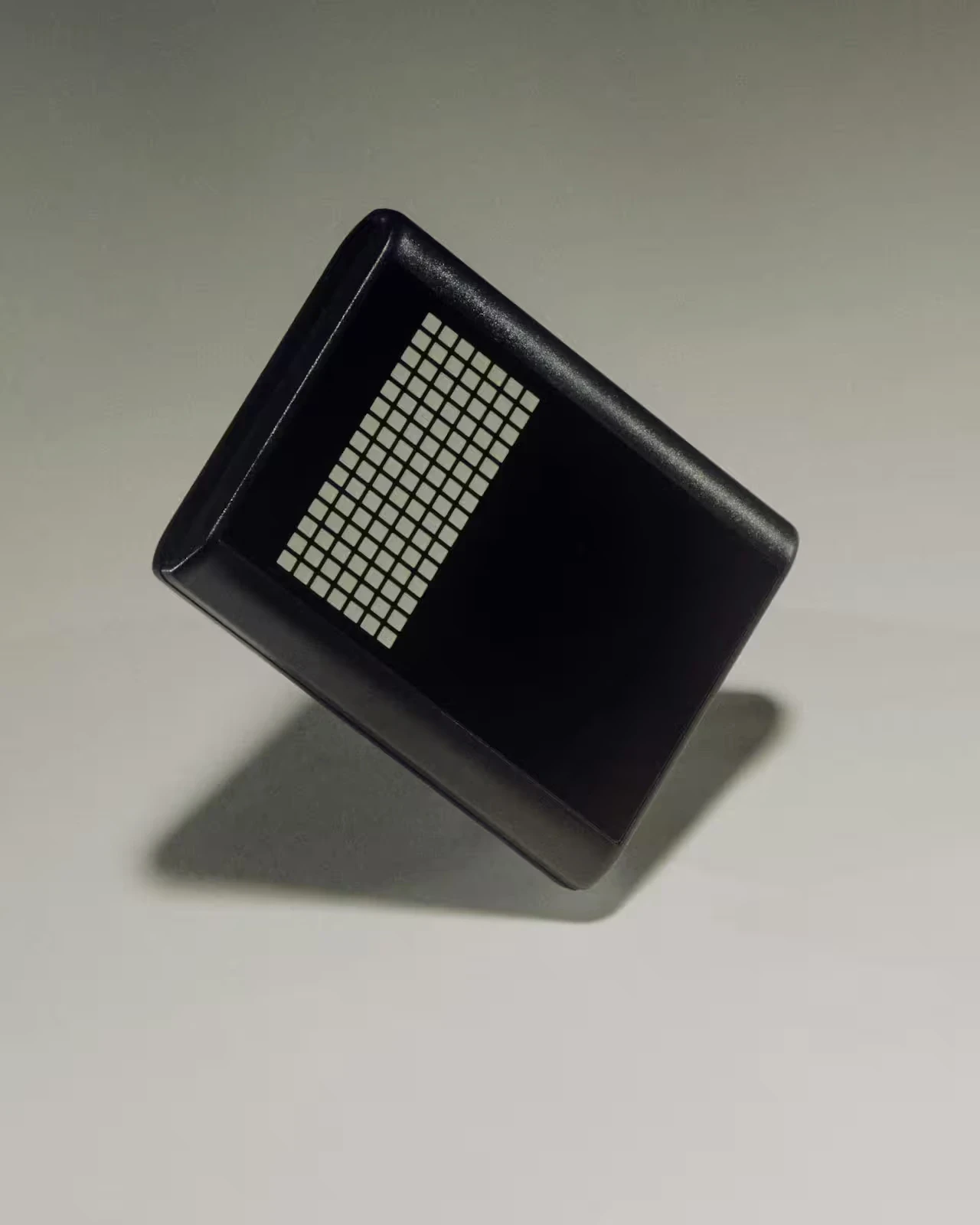 A small black rectangular device with a grid of white dots stands balanced on its corner against a neutral background.