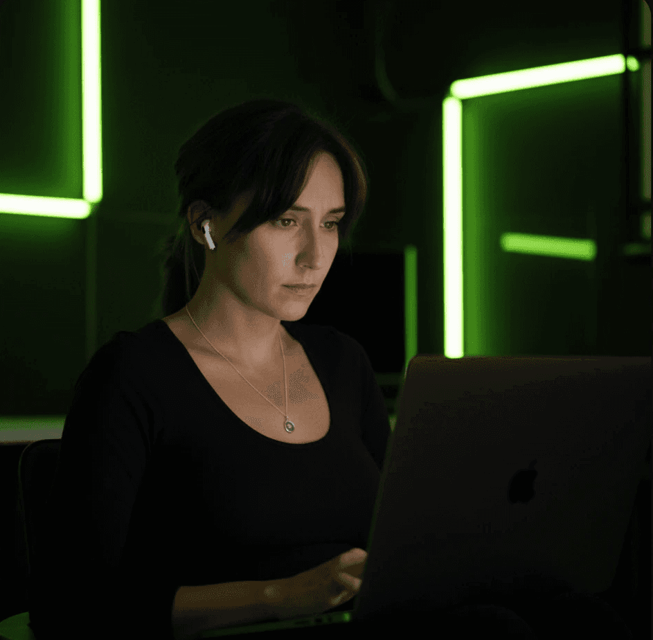Woman working on a laptop in a dark room with green neon lights in the background.