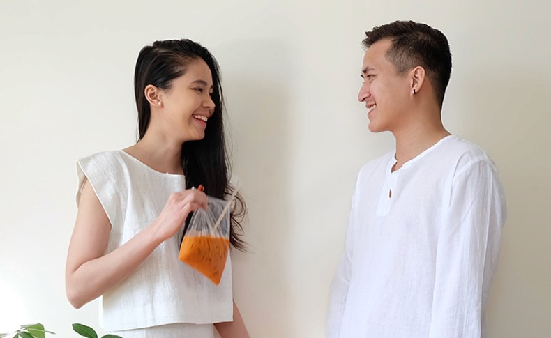 A man and a woman are standing facing each other against a plain off-white wall. The woman, on the left, has long dark hair and is wearing a white sleeveless top. She is holding a clear plastic bag containing an orange-colored substance and appears to be smiling and looking at the man. The man, on the right, has short dark hair and is wearing a white long-sleeved shirt. He also seems to be smiling and looking at the woman. A small portion of a green plant is visible in the bottom left corner of the image.