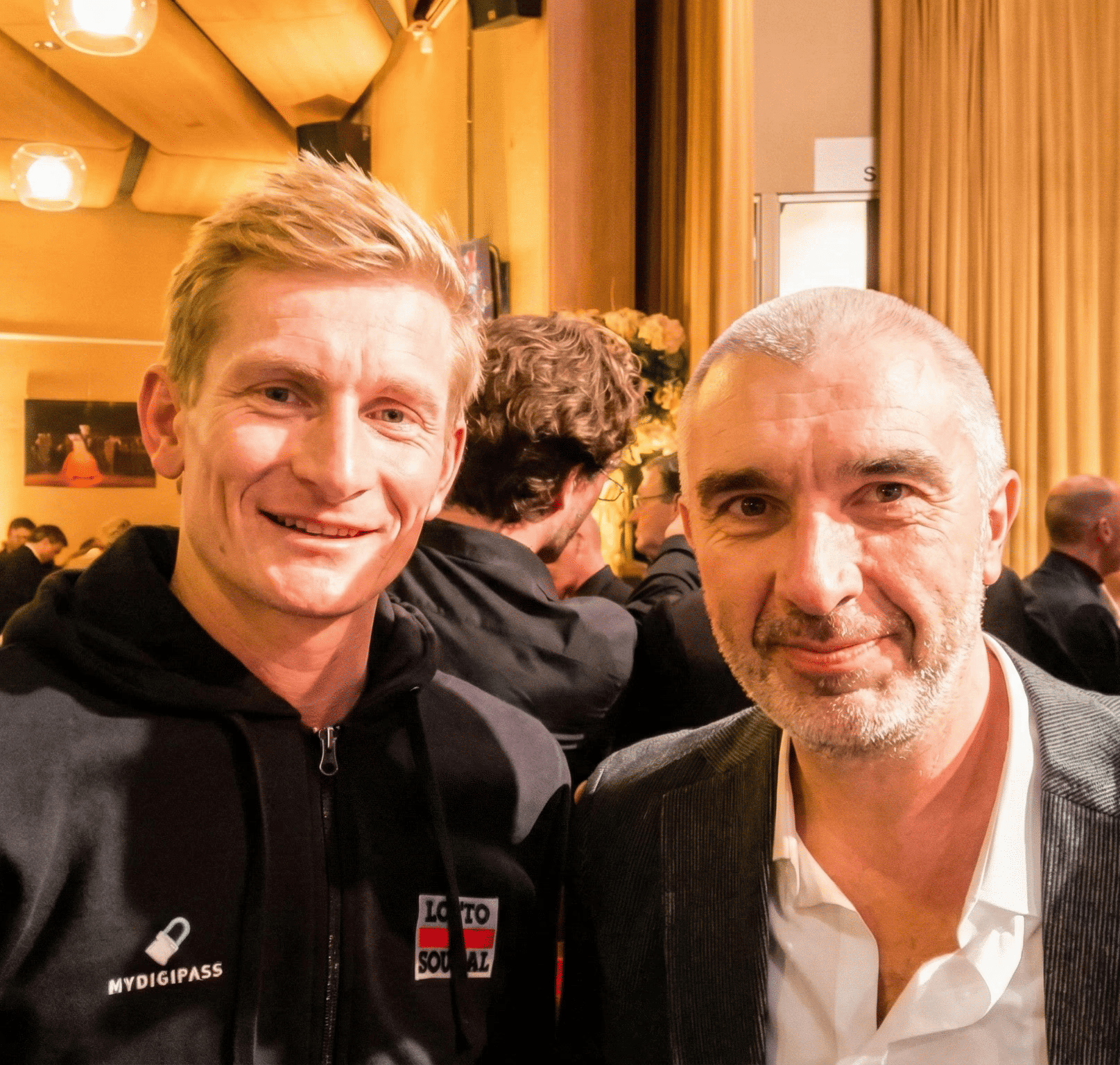 Photo du propriétaire du magasin de vélo BFZ Eghezee posant en souriant aux côtés de l'ancien cycliste professionnel André Greipel lors d'une soirée événementielle.