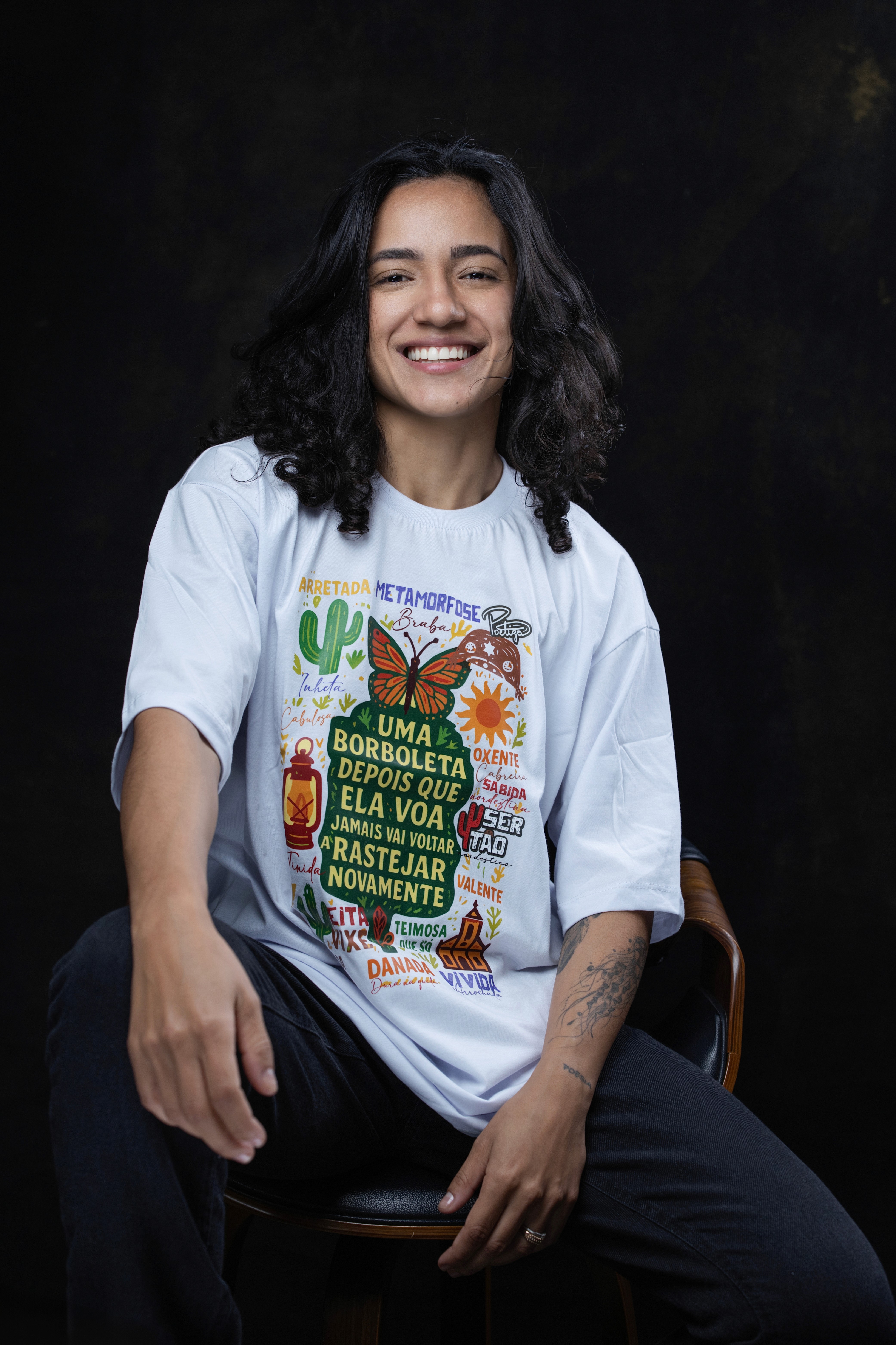 Una persona con una camiseta negra y pantalones de color claro está de pie en la cima rocosa de una montaña al atardecer, sosteniendo una cámara frente a su cara para tomar una foto. Un mar de nubes se extiende detrás de ellos, iluminado por la luz dorada del sol poniente.