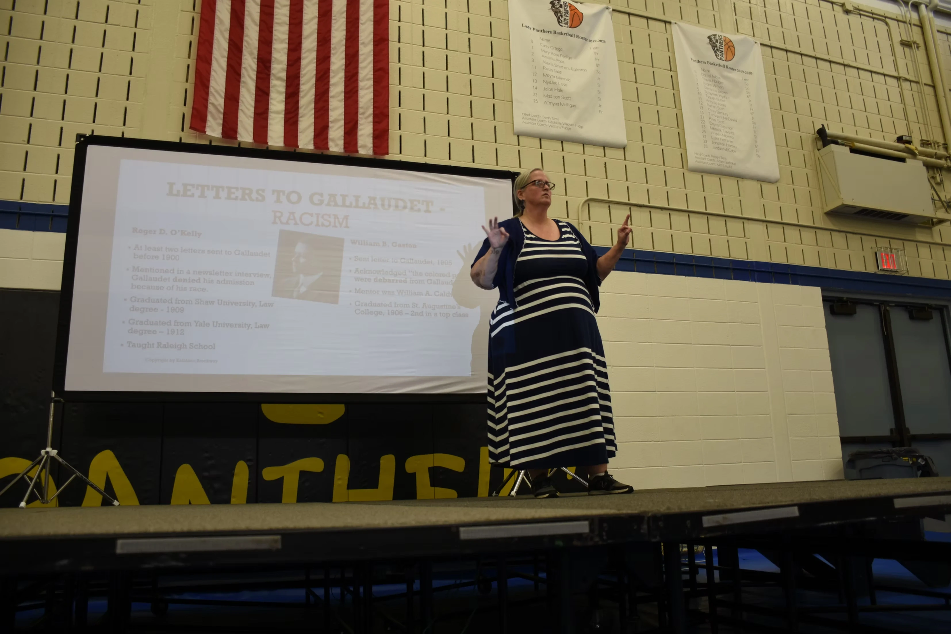 A woman in a striped dress gives a presentation on uprooting racism, gesturing with a pointer towards a projector screen.