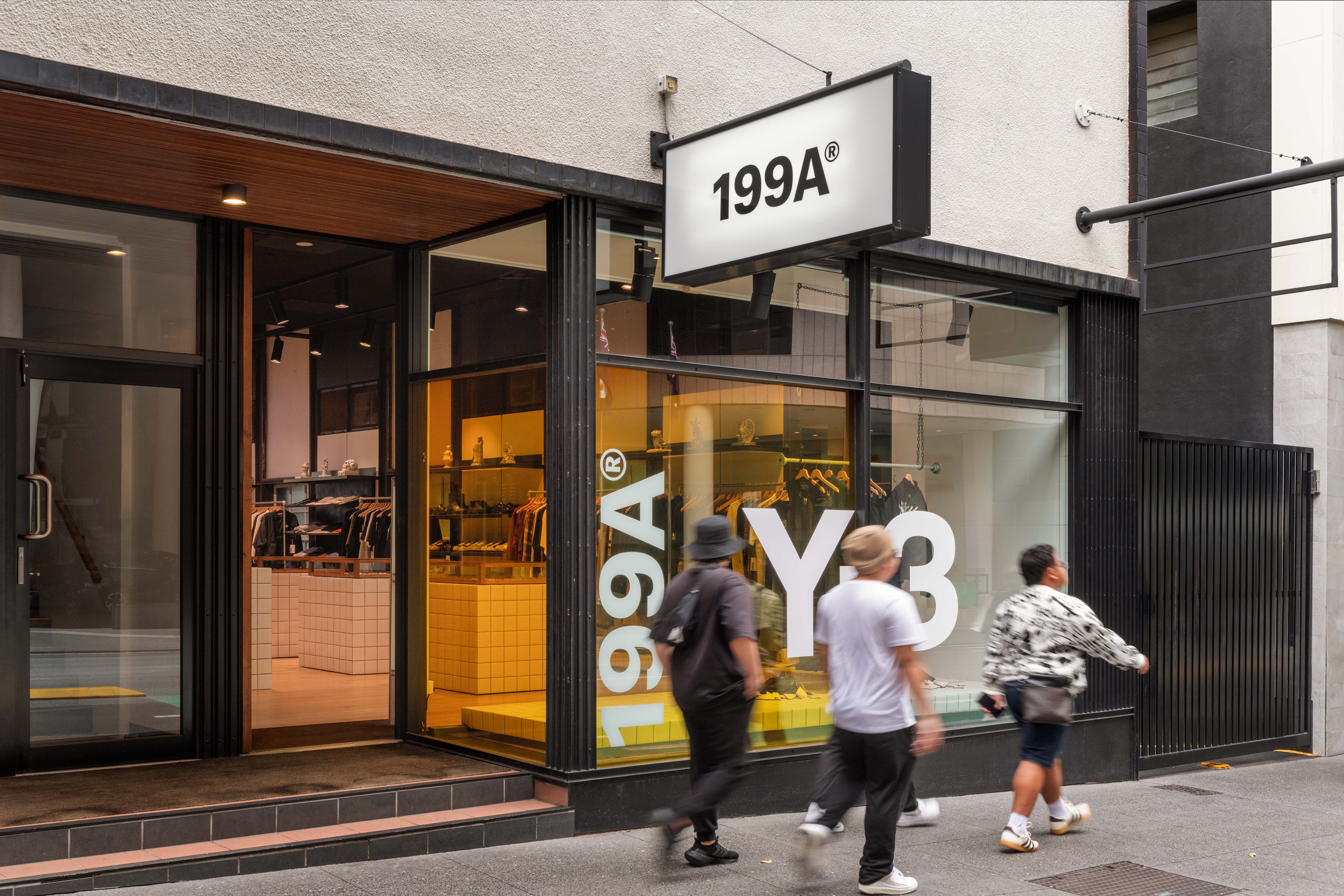 Street view of the 199A® storefront with bold window graphics and warm-lit interior visible inside, as pedestrians pass by and the space engages the surrounding city.
