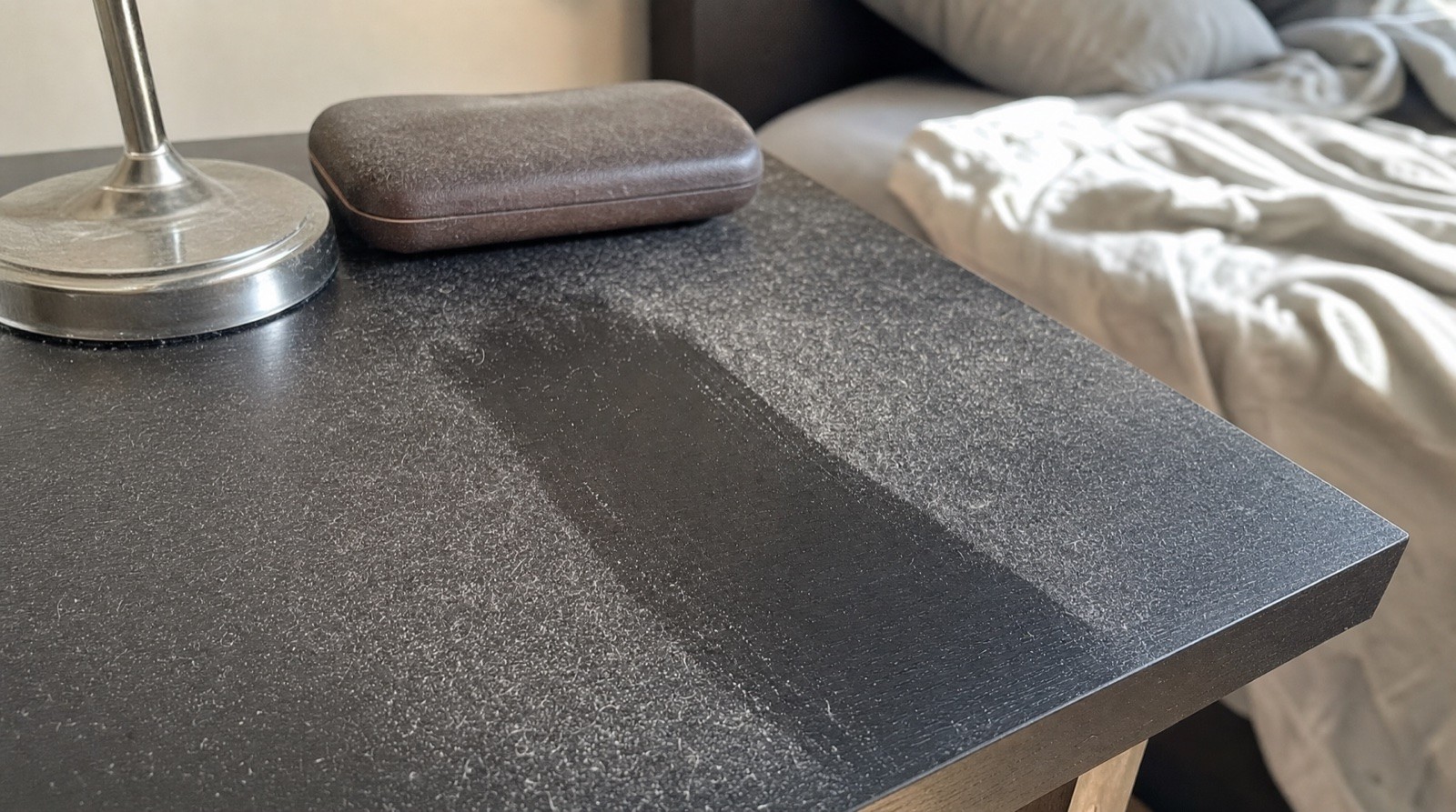 A close-up of a dusty nightstand next to an unmade bed in morning light.