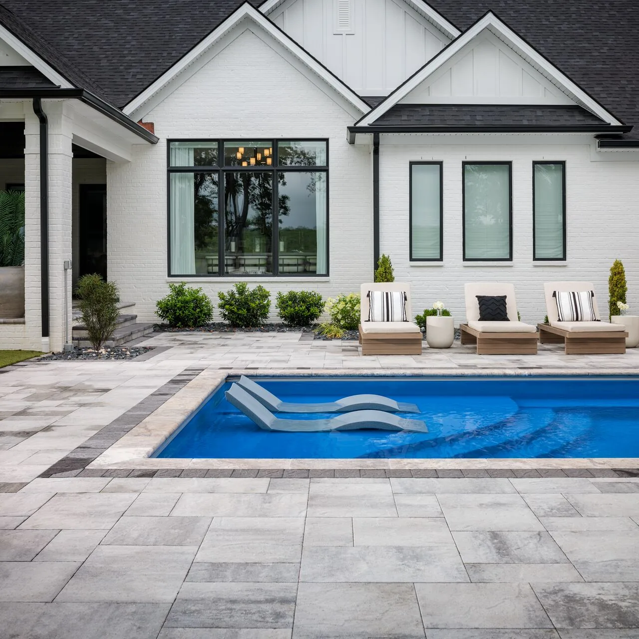 The house and pool together Pale paver pool deck outside a white house with tall windows and lounge chairs.