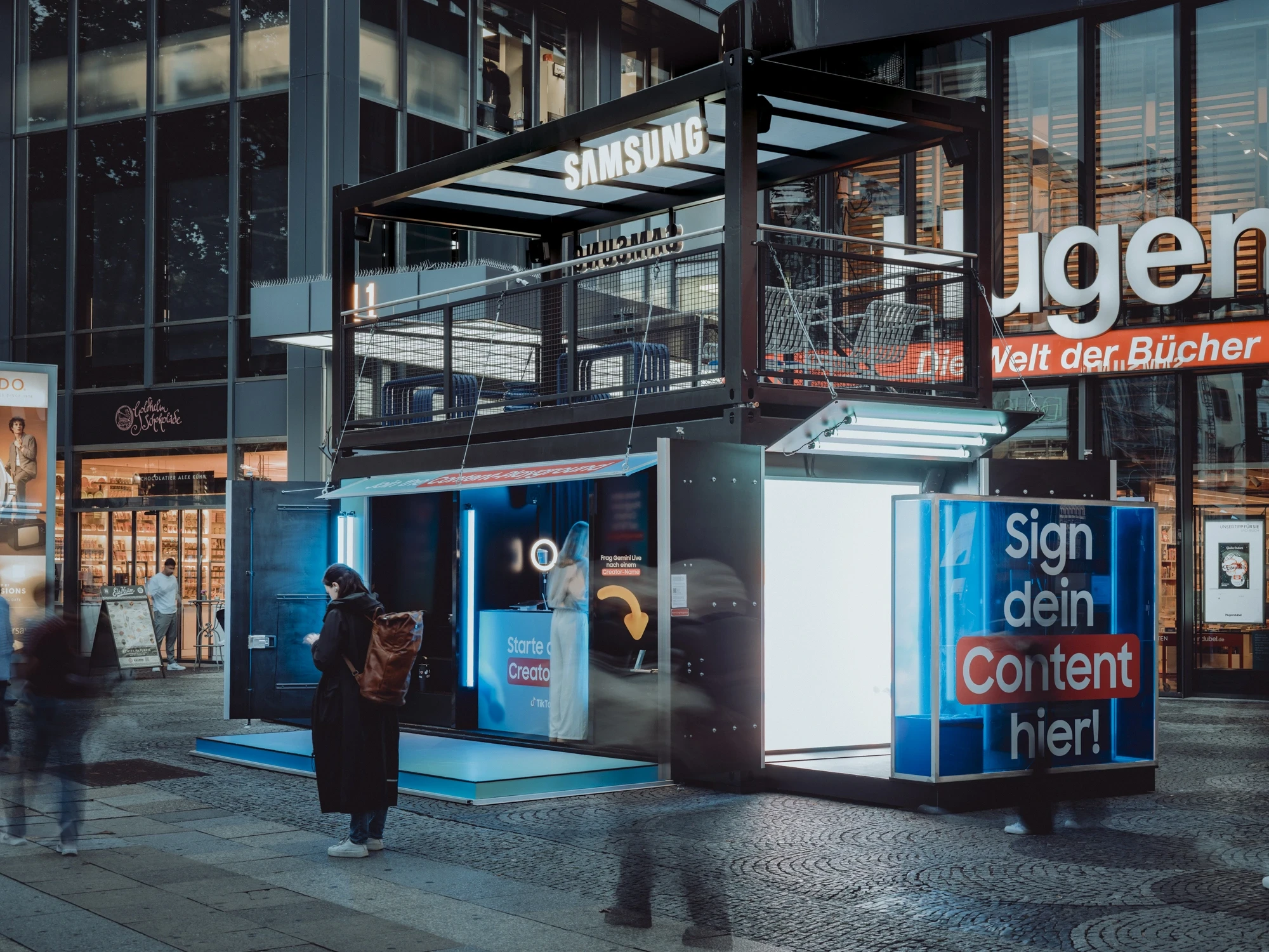 SAMSUNG x TIKTOK container module at night in a shopping street with open blue-lit experience rooms, roof deck and illuminated campaign graphics.